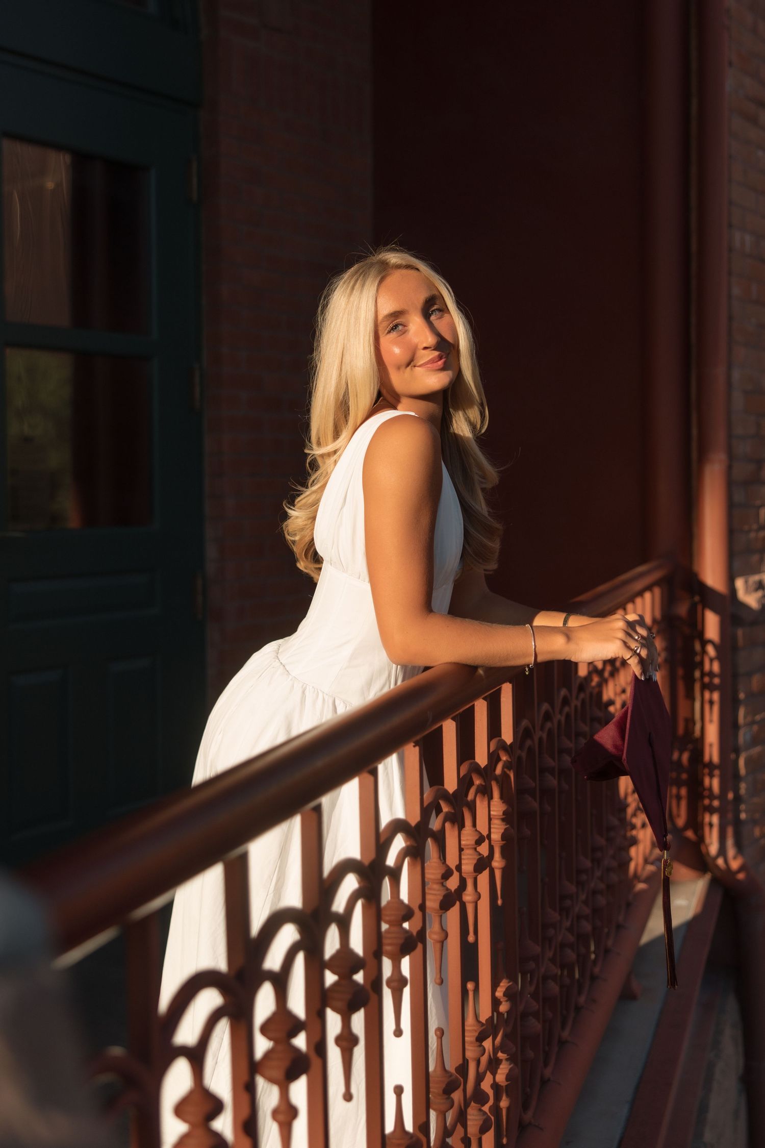Blonde woman in white dress smiling on ornate iron balcony railing in golden sunset light.