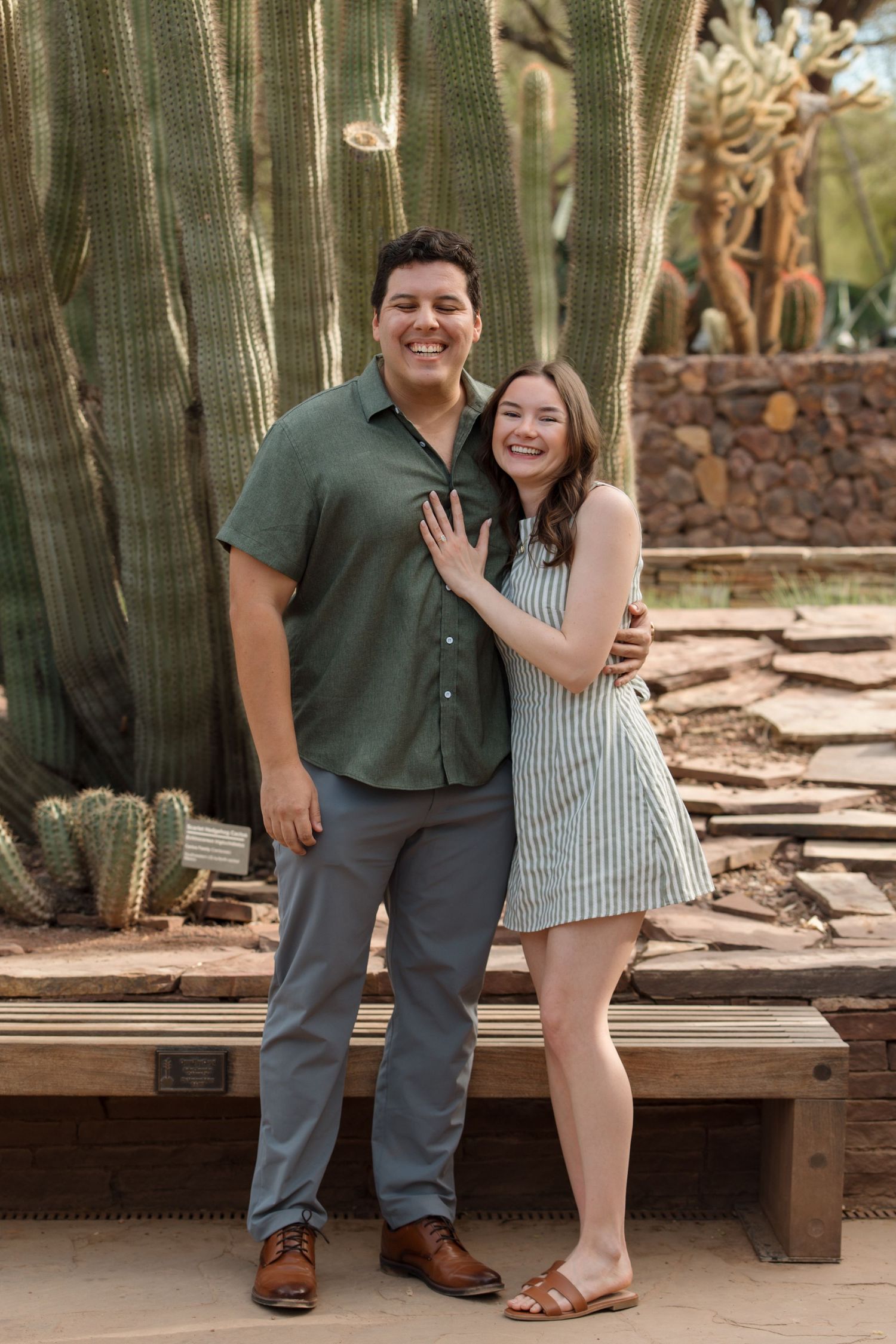 Happy engaged couple posing together in front of tall cacti, woman showing off engagement ring.