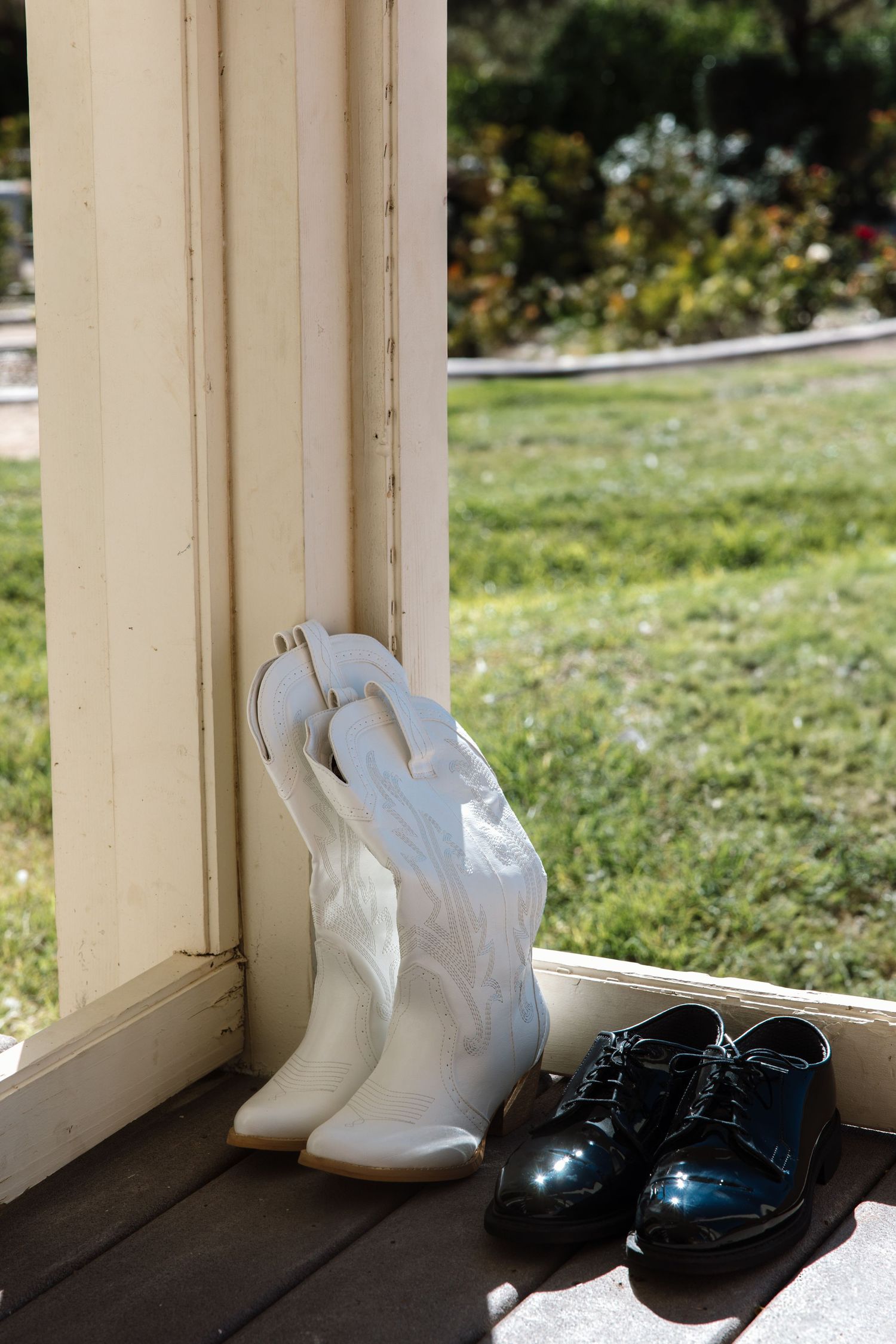White cowboy boots and black patent leather dress shoes sit side by side on a sunny porch at a wedding.