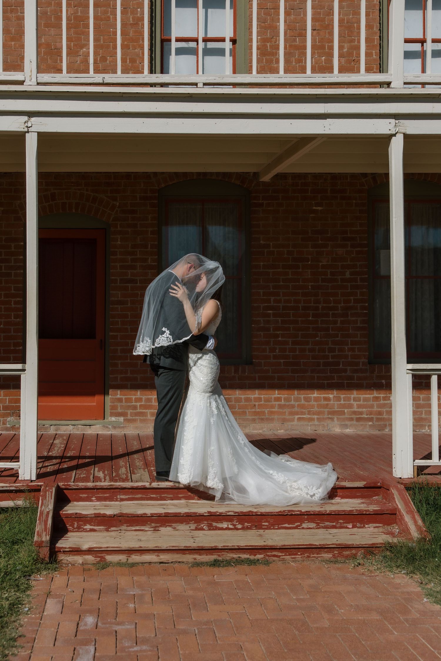 Bride and groom share a kiss under a veil on steps of a brick building.