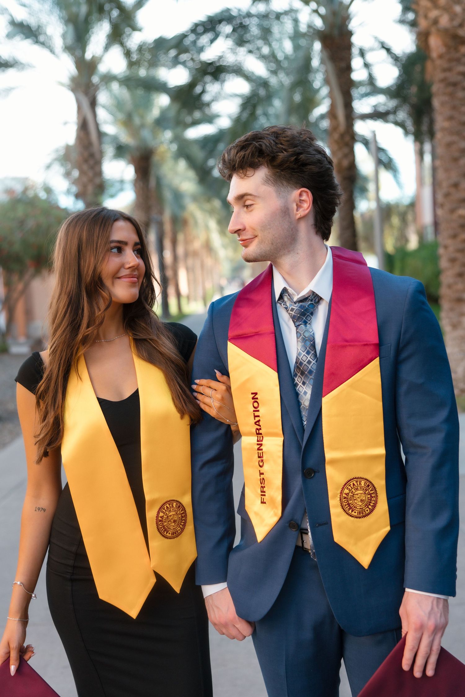 Two college graduates wearing gold graduation stoles walk together outdoors among palm trees.