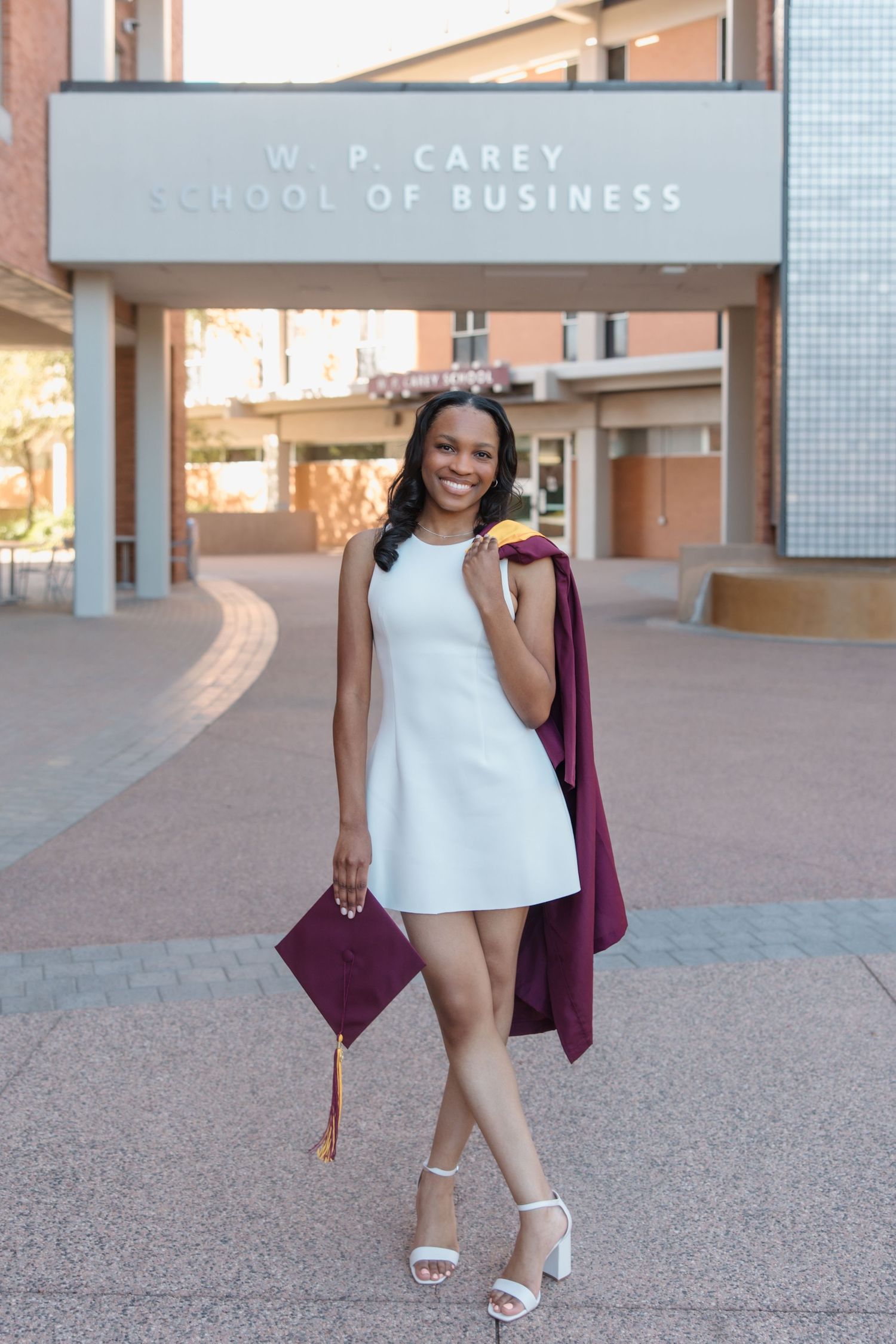 Smiling graduate in white dress holds maroon cap and gown outside W.P. Carey School of Business building.