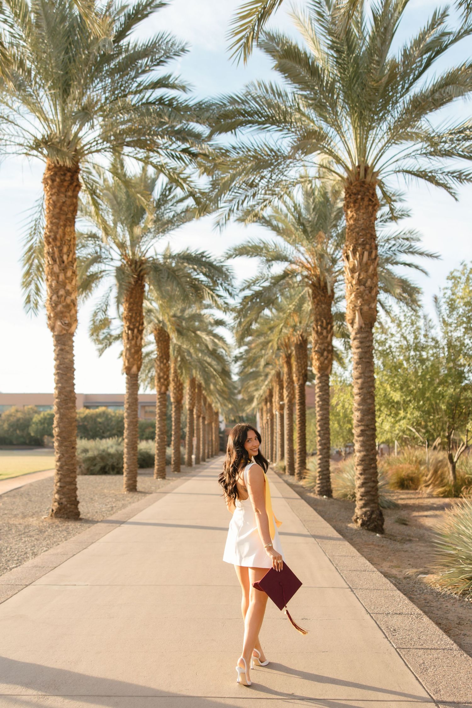 Young woman in white dress holding burgundy graduation cap walks along palm tree-lined path in sunny desert setting.