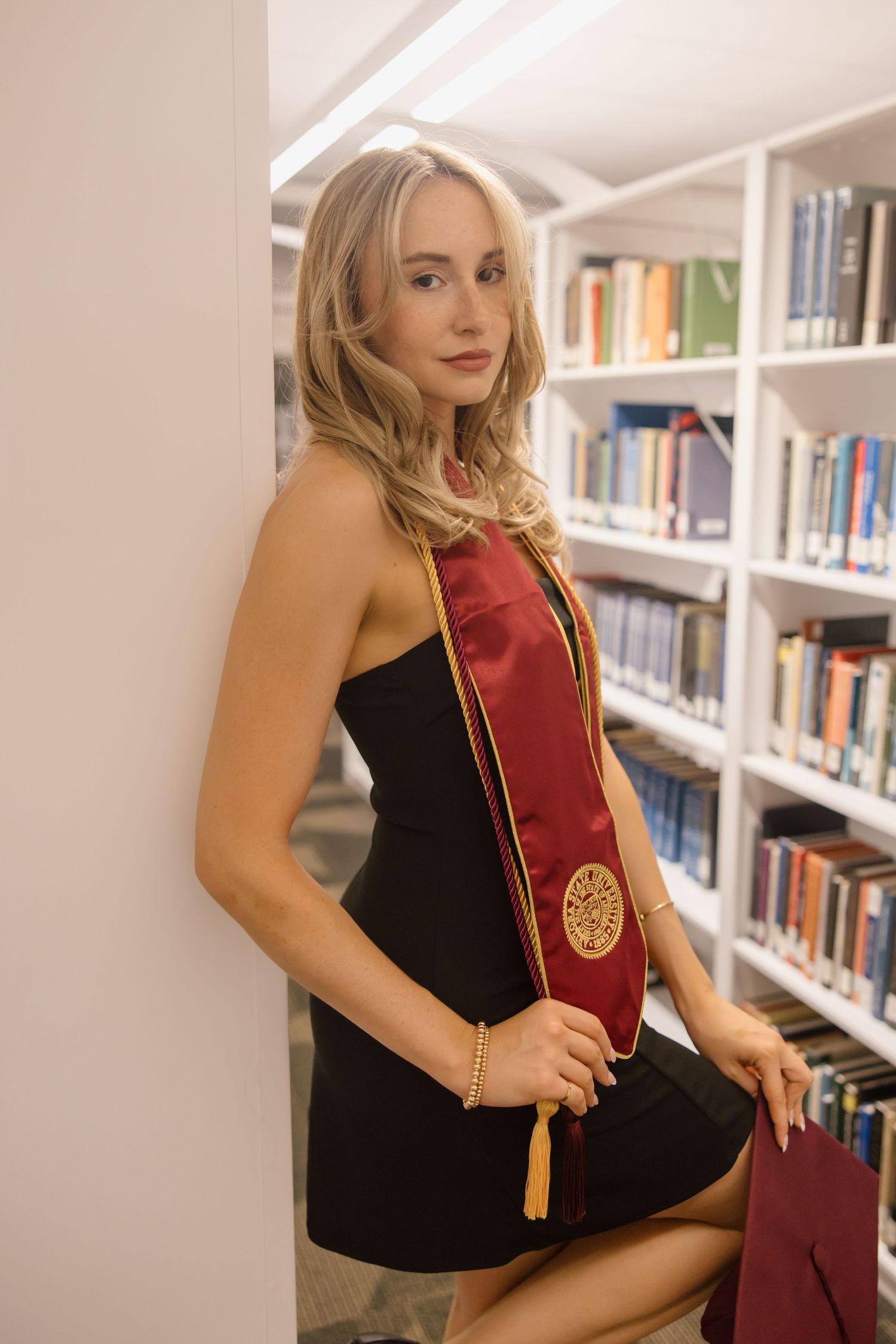 Young blonde woman in black dress wearing maroon graduation stole poses in library for graduation photo.