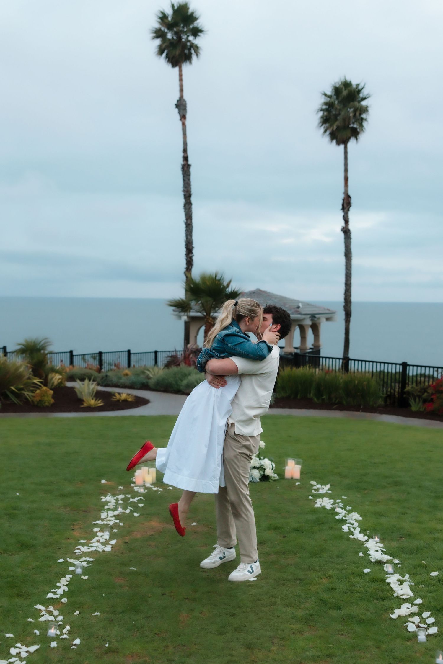 Man lifts and kisses his newly engaged partner on lawn with heart-shaped rose petal path by ocean.