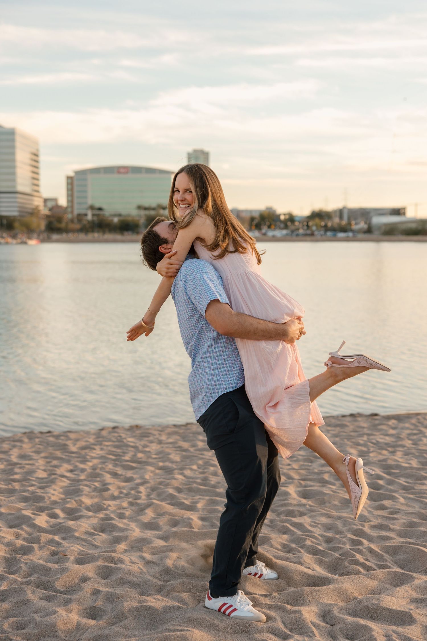 Couple laughing joyfully on sandy beach at sunset, man lifting woman in pink dress near waterfront.