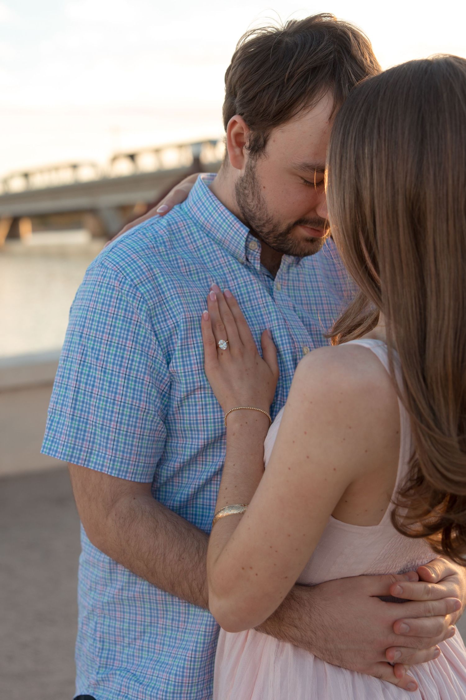 Engaged couple embraces closely outdoors at sunset, woman displaying diamond engagement ring on her hand.