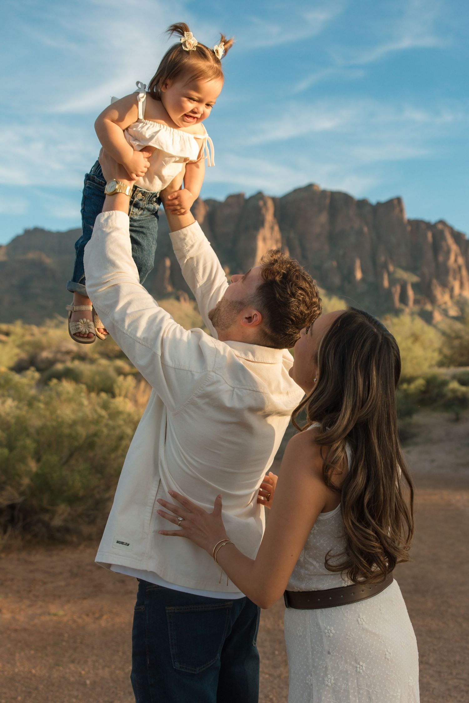 Father lifting smiling toddler girl high while mother embraces him in a desert mountain setting.