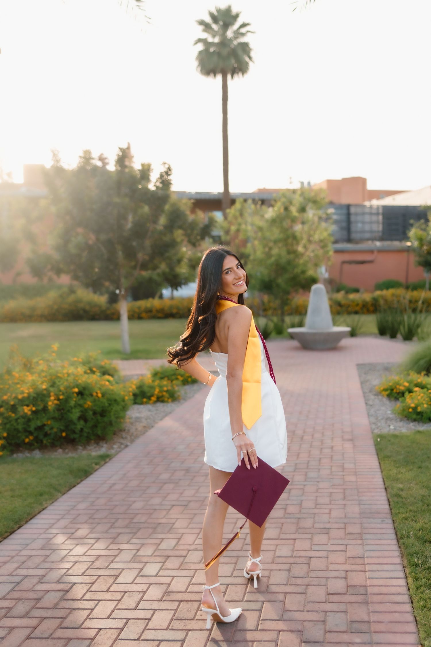 Female graduate in white dress with gold and maroon stole walking sun-drenched campus pathway.
