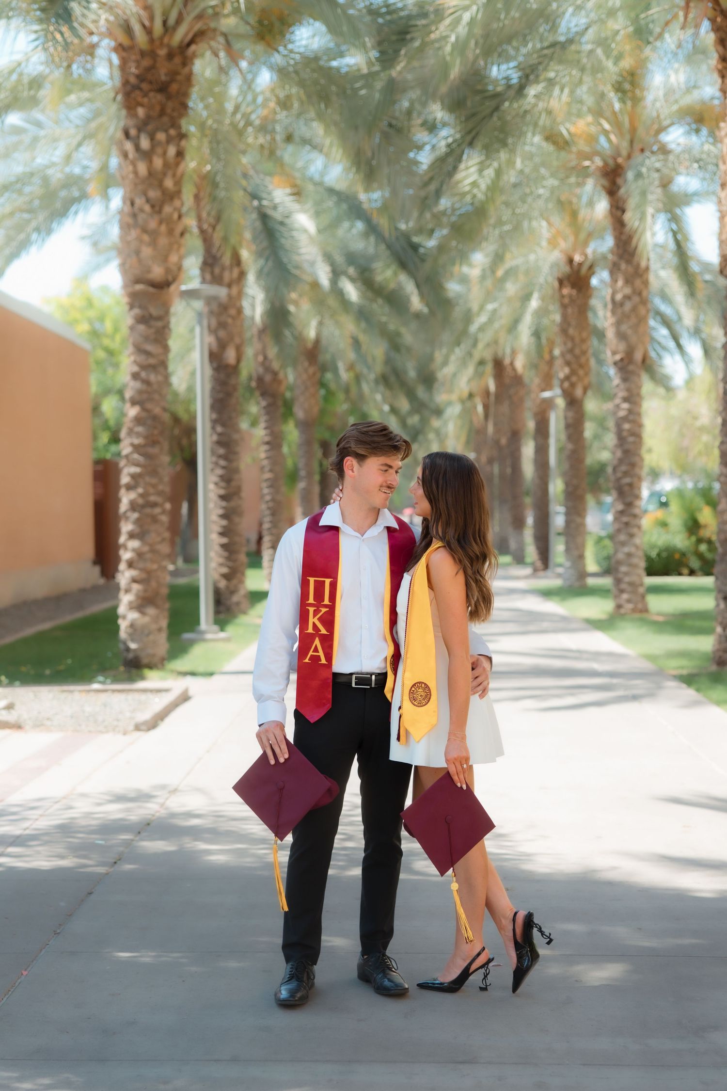 Graduating couple in stoles gazing at each other on sunny palm-lined university pathway.