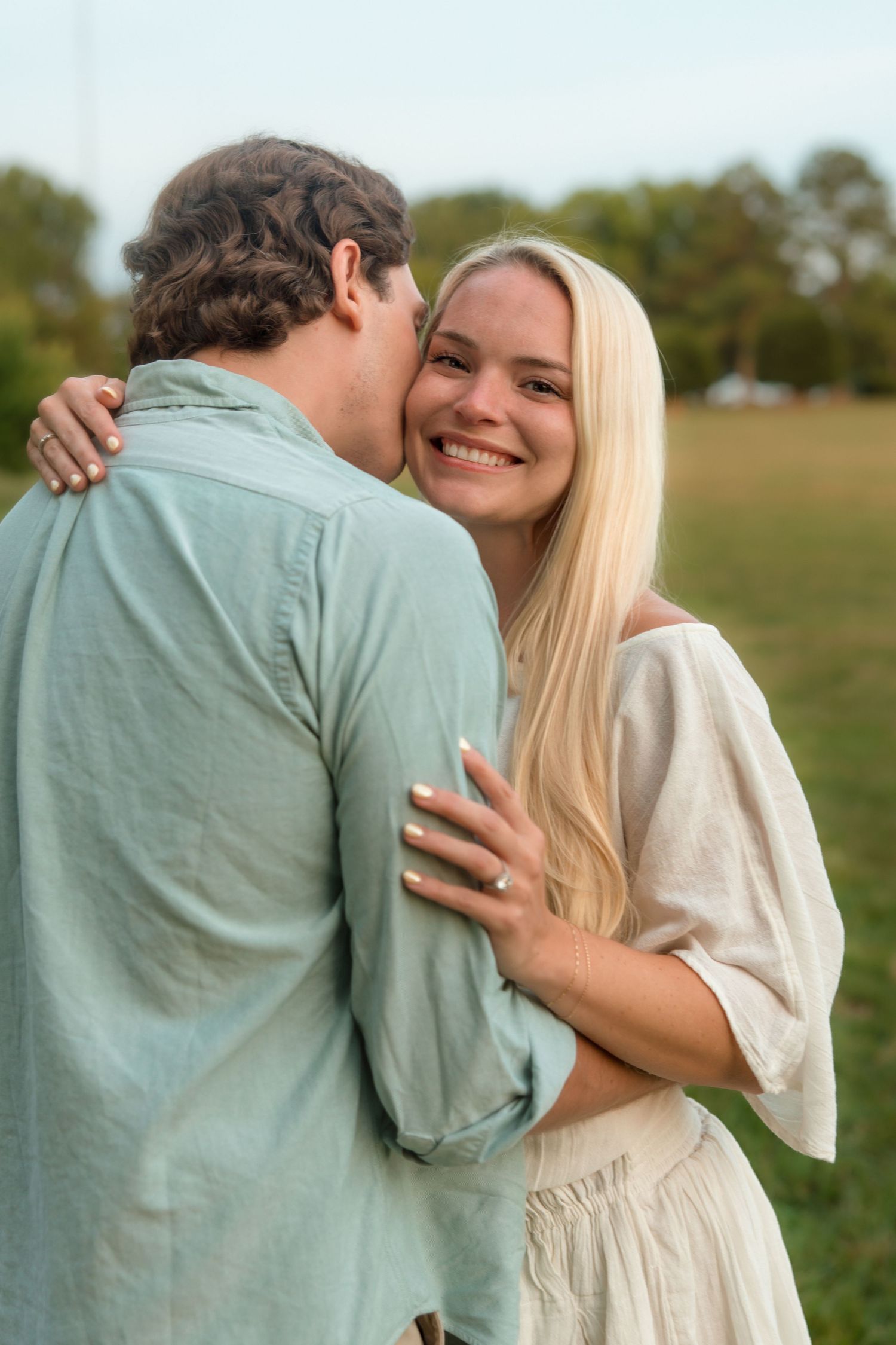 A smiling blonde woman embraces a man who kisses her cheek during an outdoor couples photo session.