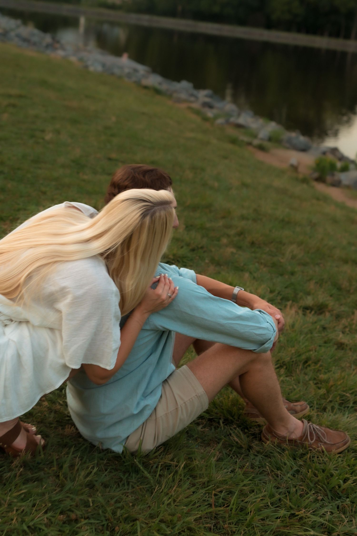 Blonde woman resting her head on her partner's shoulder while sitting by a lake.
