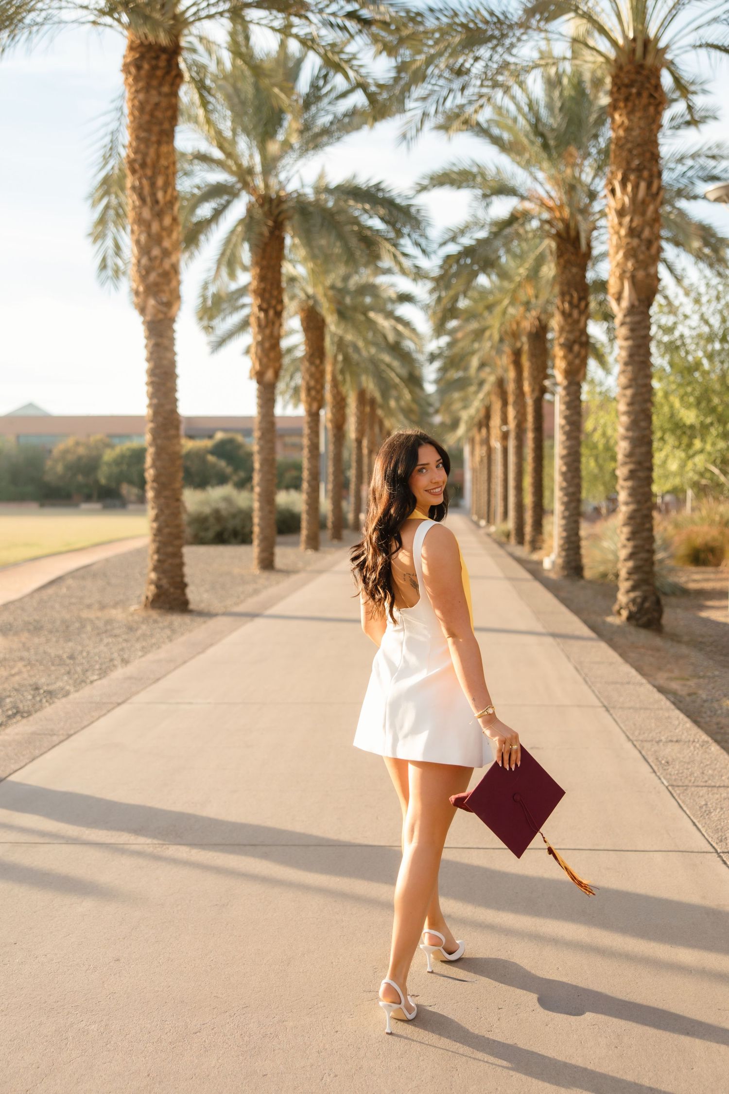 Young woman in white dress holding maroon graduation cap walks along palm tree-lined path.