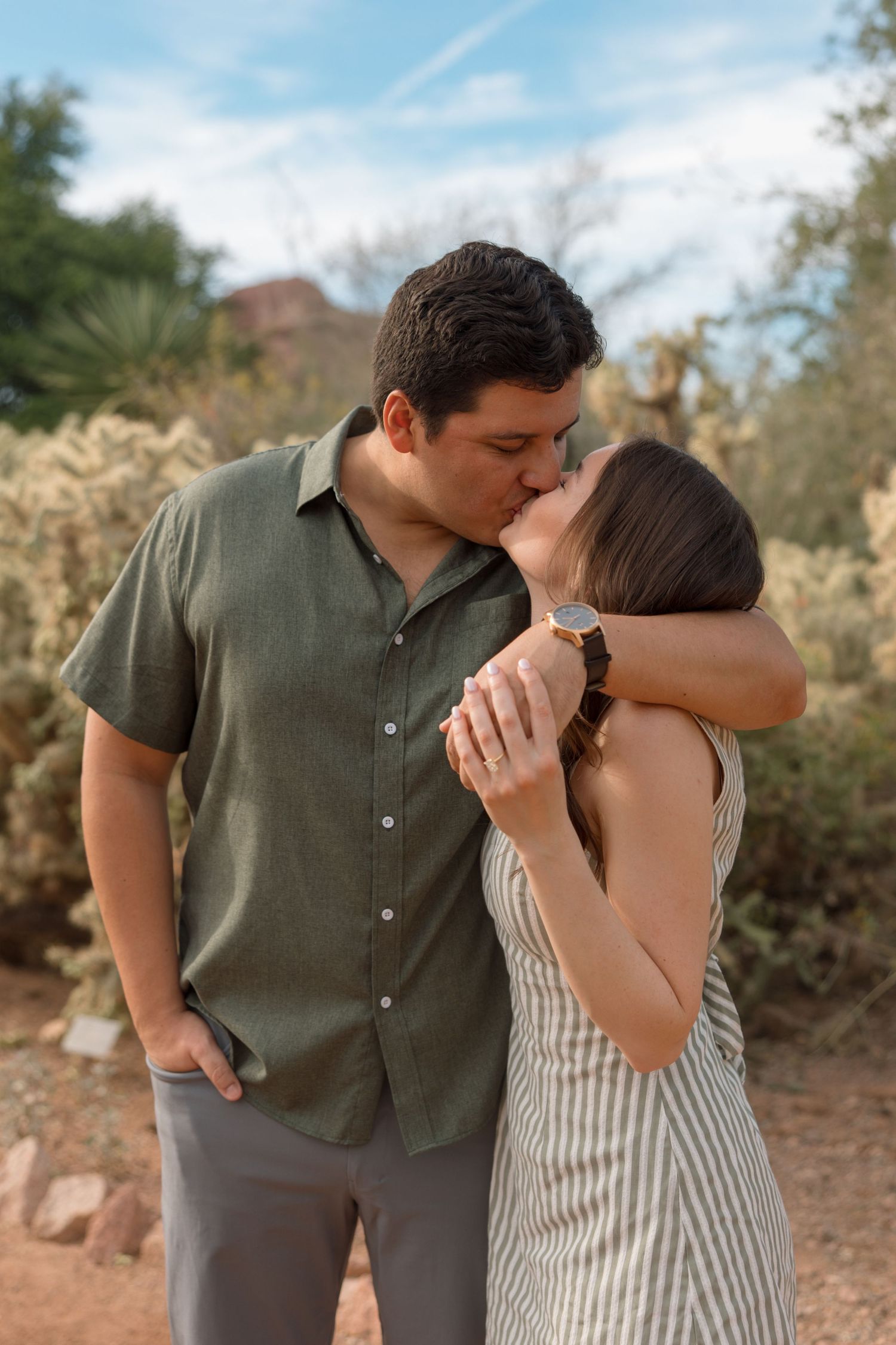 Couple sharing a romantic kiss outdoors in a desert garden setting, woman in striped dress holding mans hand.