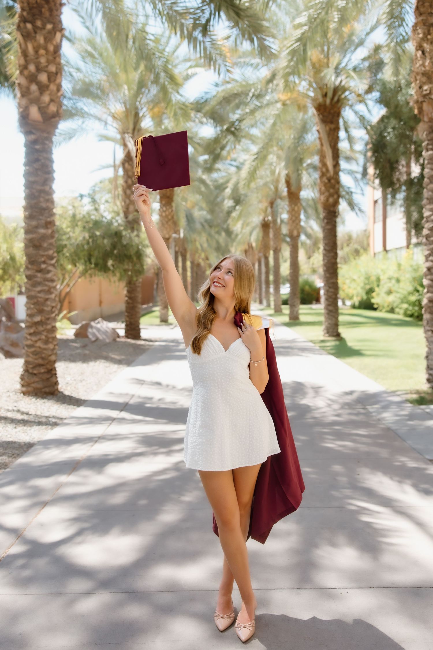 Young woman in white dress celebrates graduation, holding cap and maroon gown among palm trees on a sunny campus.
