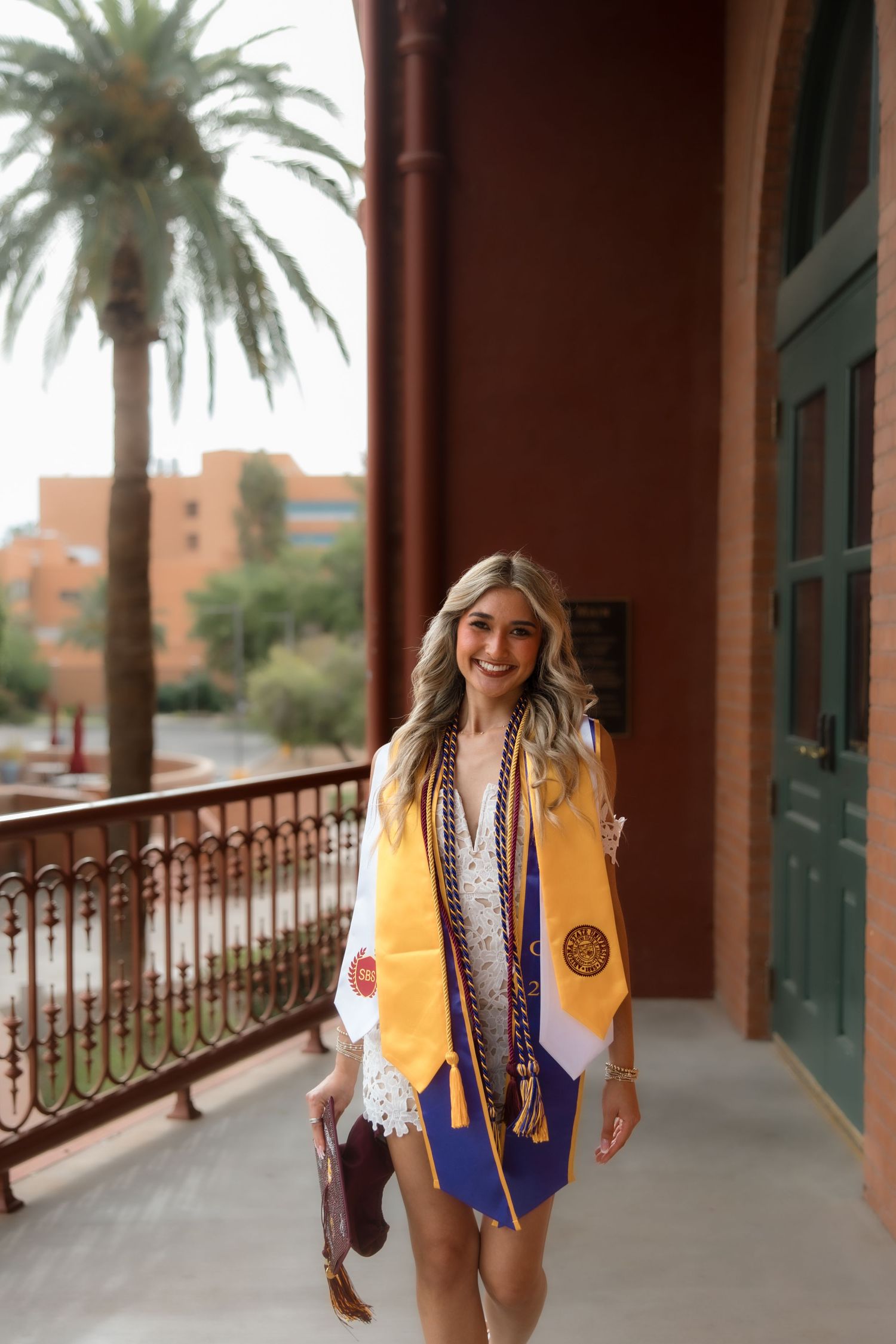 Smiling college graduate wearing gold stole and honor cords poses on a red building balcony with palm trees.
