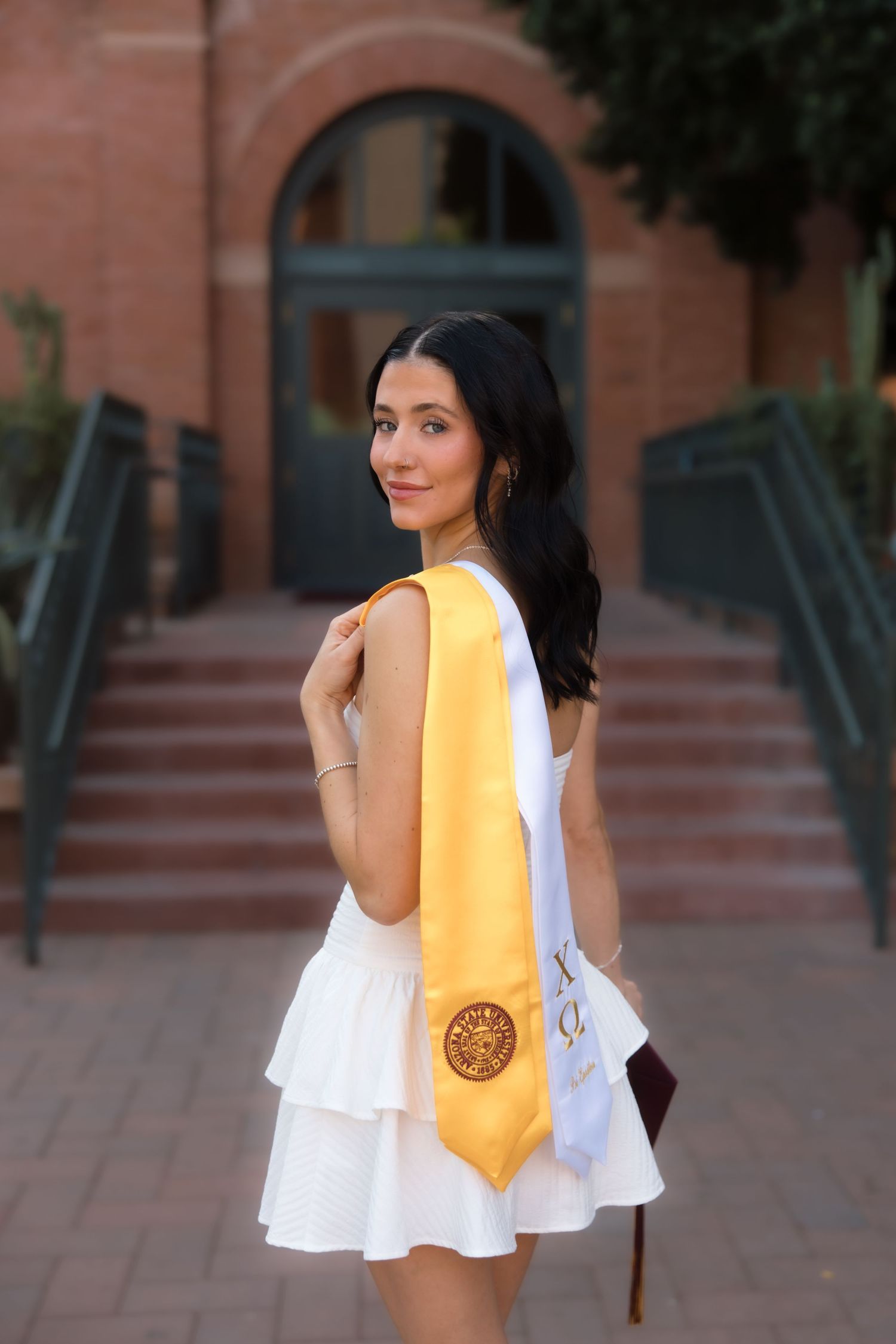 Young woman in white dress and gold graduation stole poses outdoors near brick building steps.