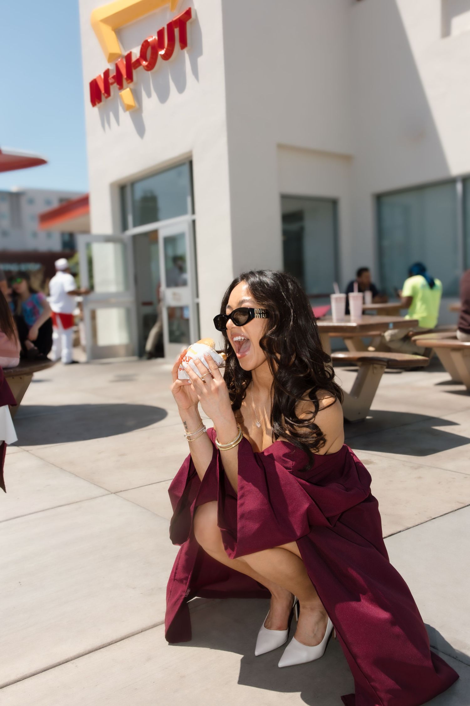 Female graduate in burgundy gown and sunglasses excitedly holds an In-N-Out burger outside the restaurant.