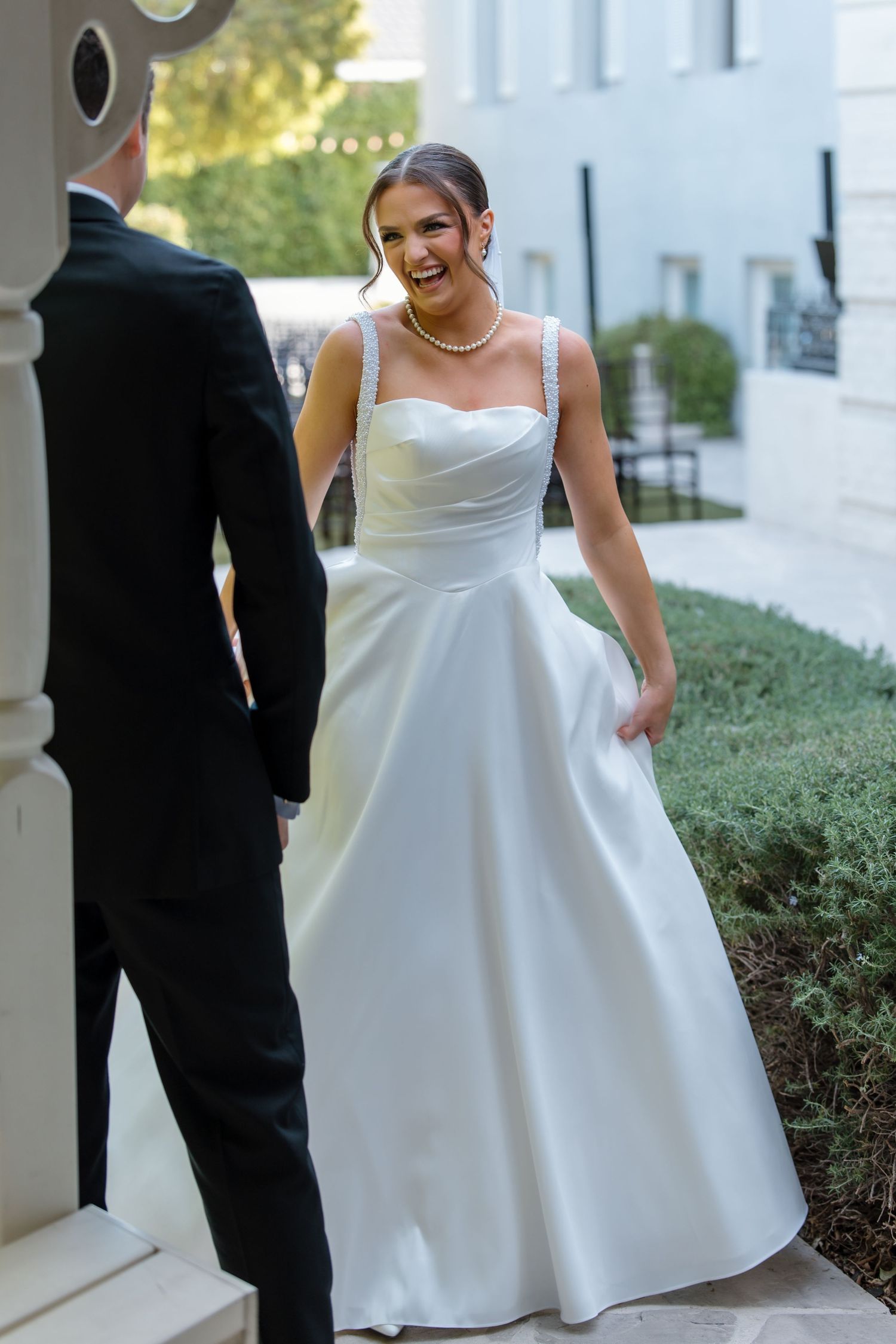 Radiant bride in white satin A-line gown with pearl necklace laughs during first look with groom.