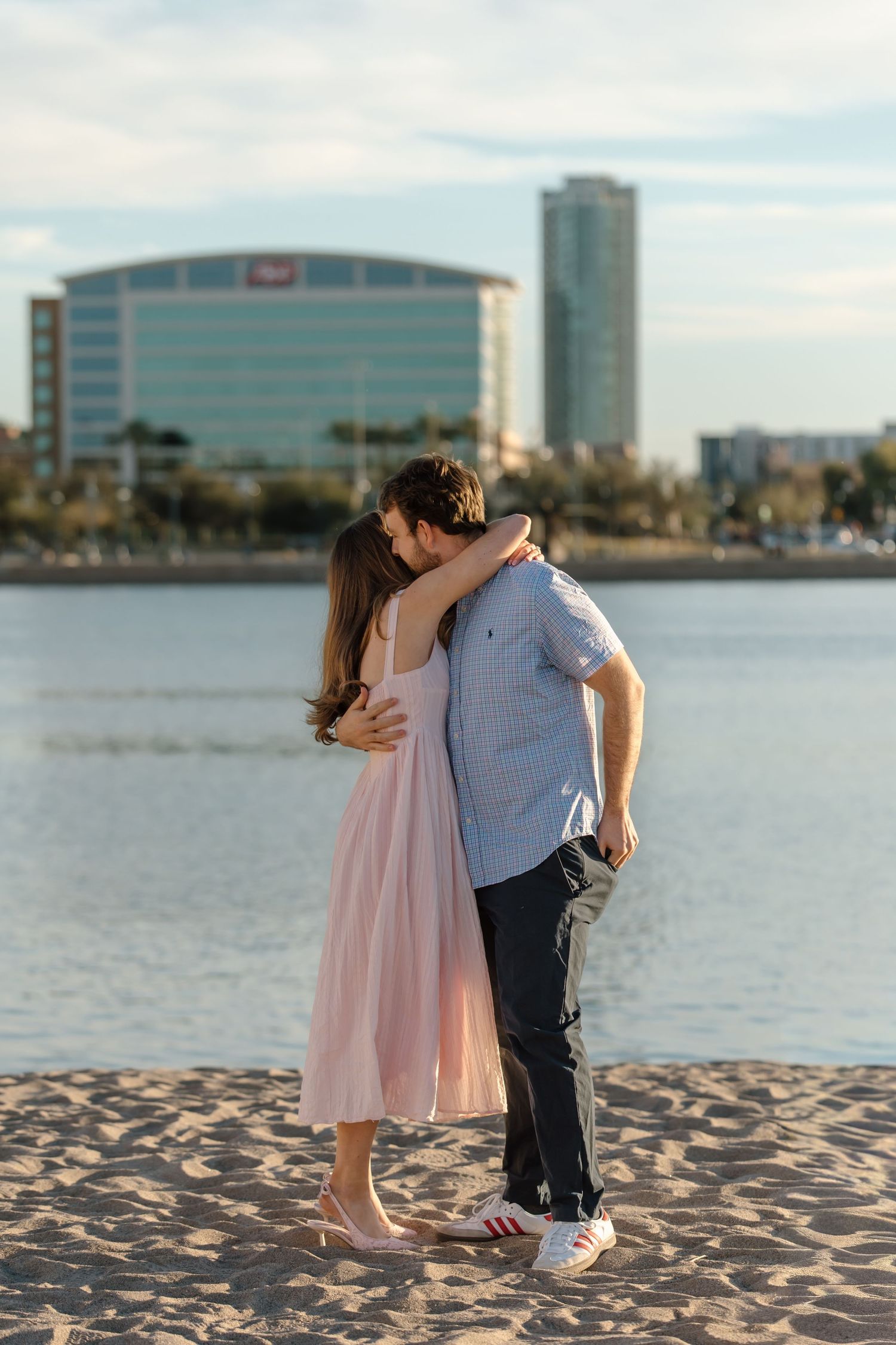 Couple embracing on sandy beach with city skyline and waterfront buildings in the background on a sunny day.