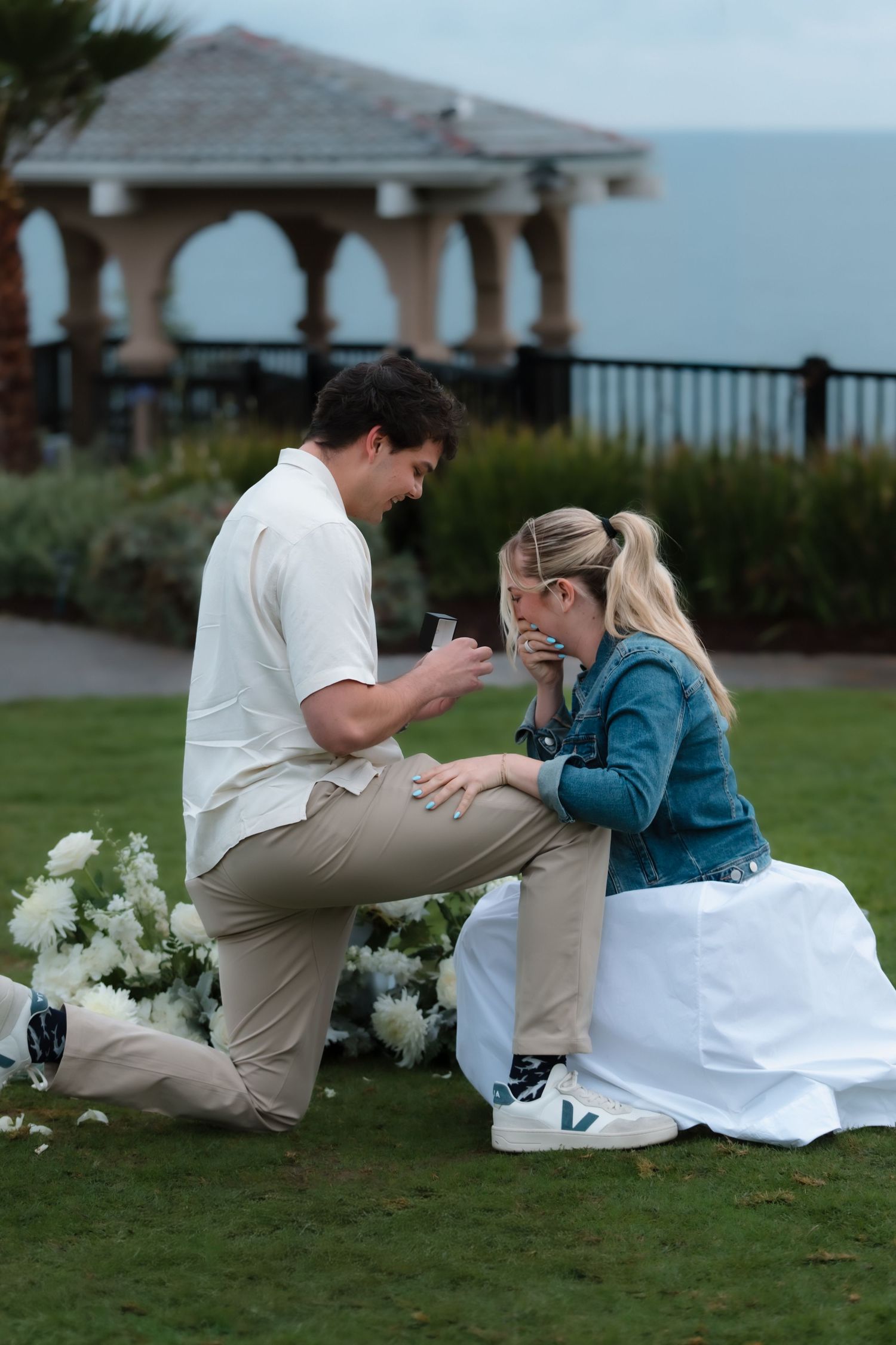 Emotional marriage proposal on lawn as man kneels showing ring box to tearful blonde woman.