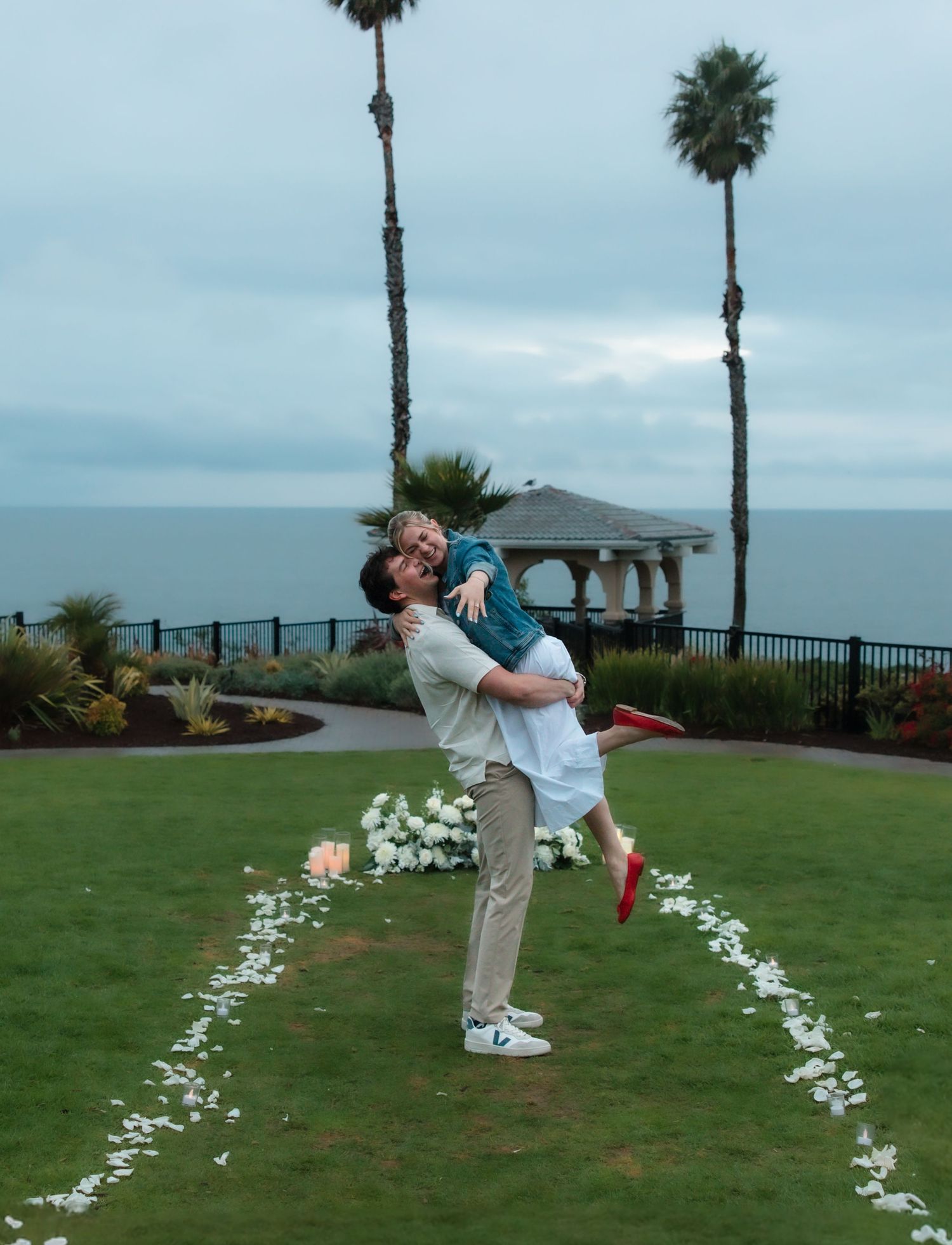 Joyful man lifting woman during outdoor engagement celebration near ocean gazebo.