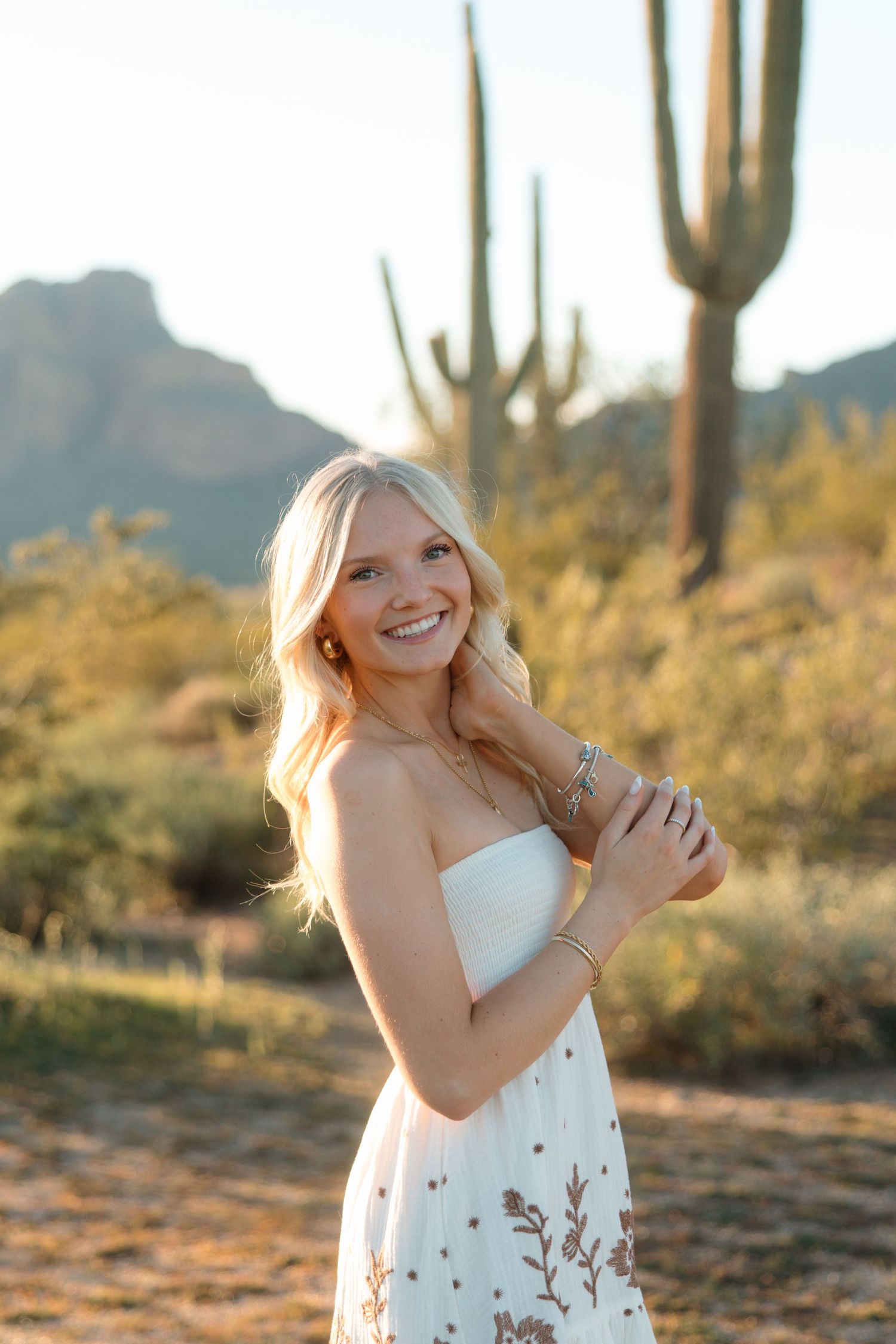 Blonde woman in white embroidered strapless dress laughing in golden hour Arizona desert setting.