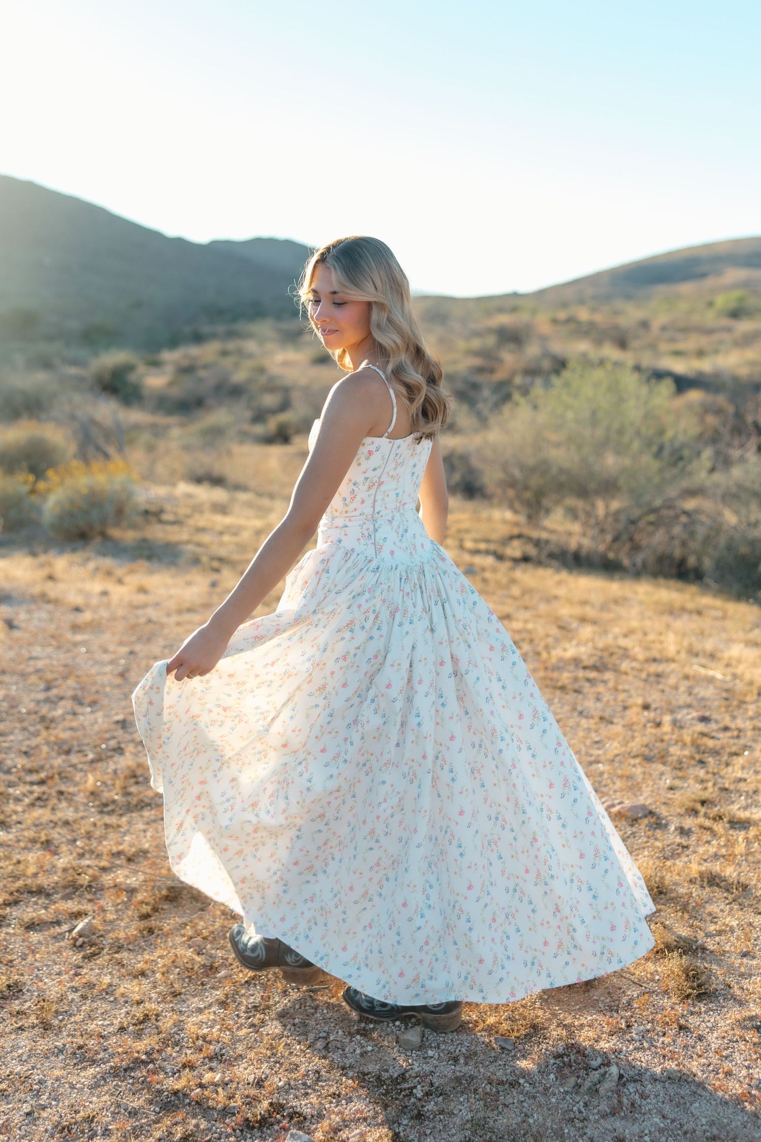 Blonde woman spinning in floral maxi dress in desert landscape at golden hour.
