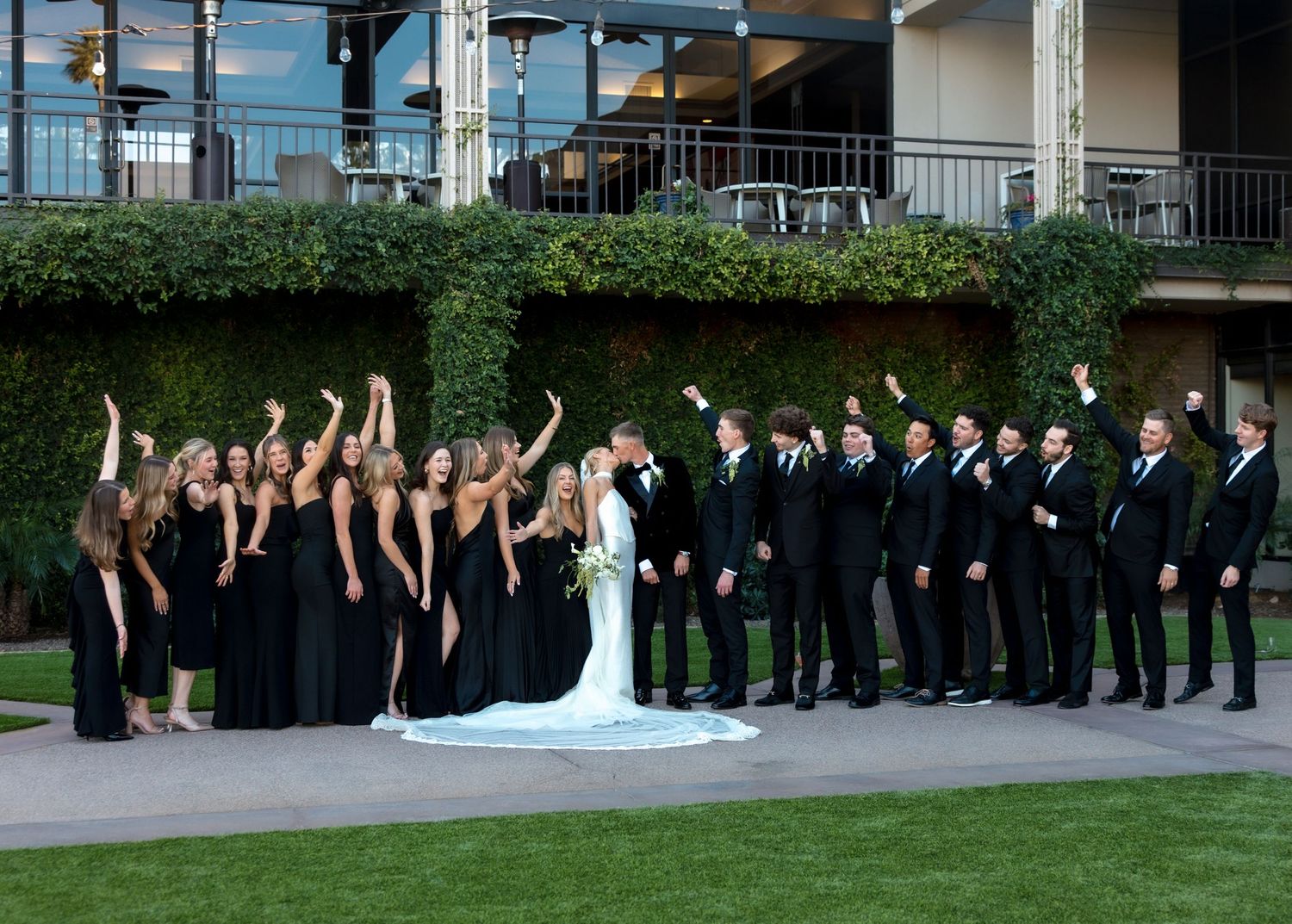 Wedding party in black attire cheers on lawn with bride and groom at elegant outdoor venue.