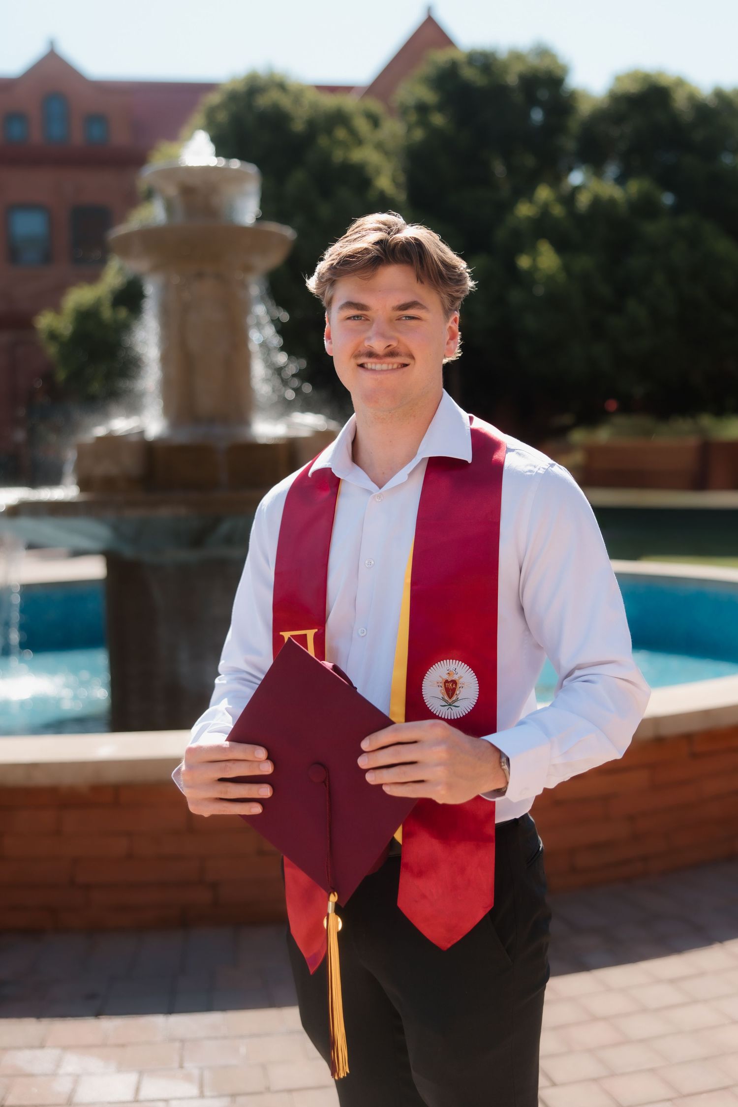 Smiling college graduate in white shirt holding maroon cap with graduation stole in front of campus fountain.