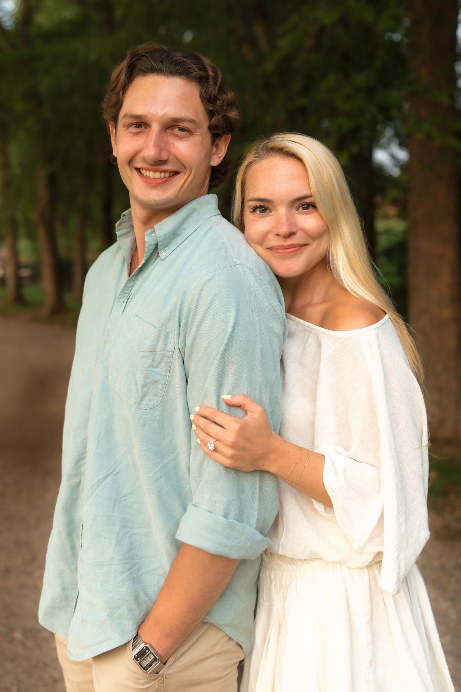 Smiling couple embracing outdoors, man in light blue shirt and woman in white dress, posing on a tree-lined path.