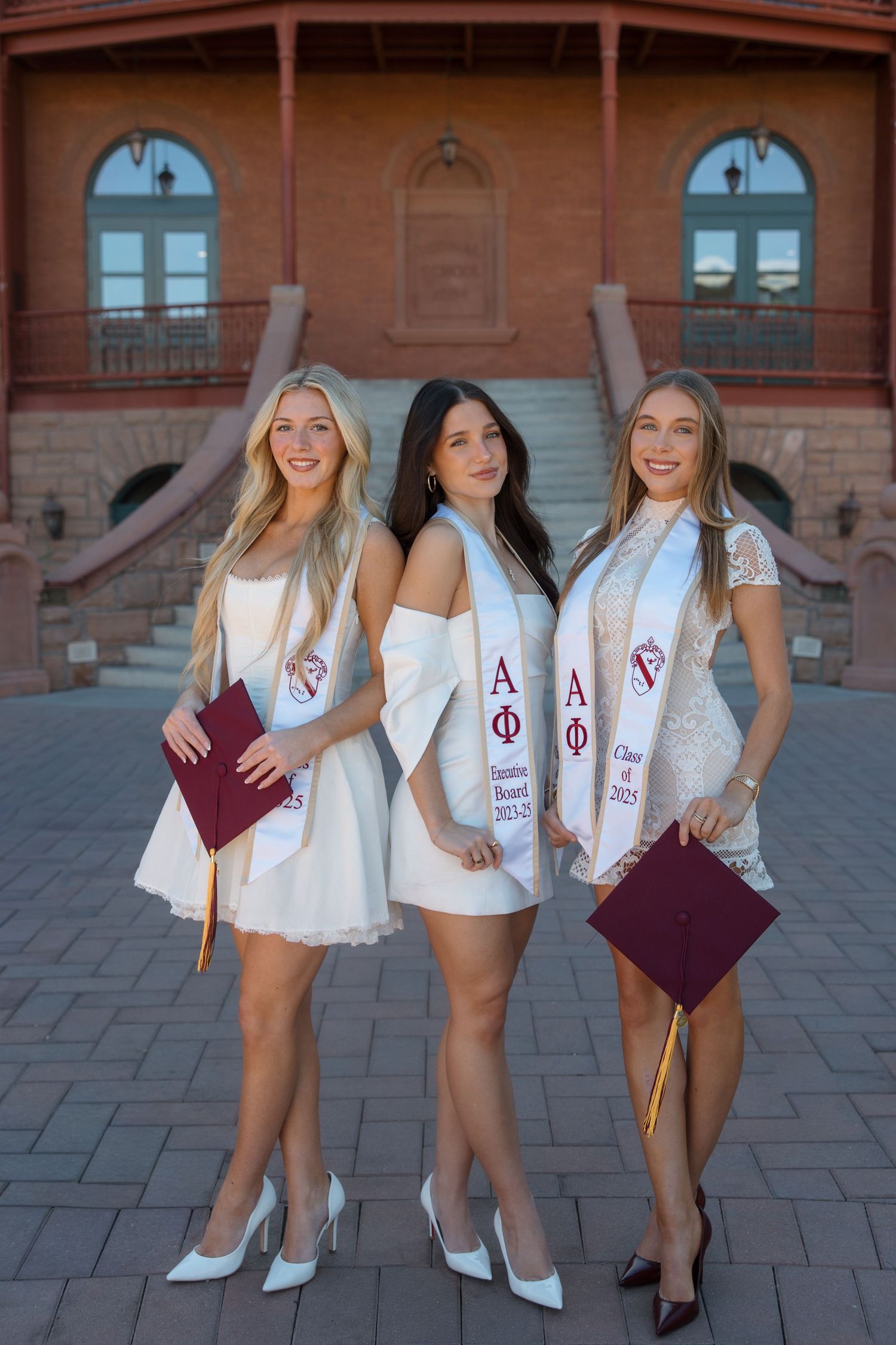 Three women in white dresses wearing Alpha Phi sorority graduation stoles pose together outdoors.