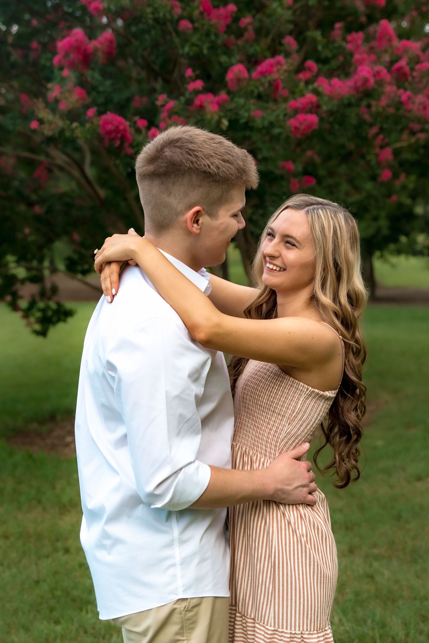 Young couple embracing outdoors near blooming pink crepe myrtle tree, smiling girl in striped dress with long wavy hair.