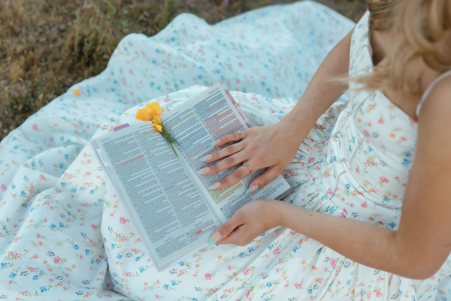 Woman in floral dress reading a book outdoors with a yellow flower as a bookmark.