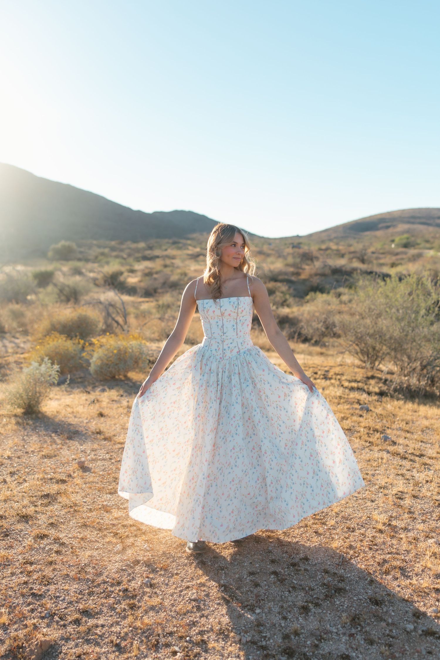 Woman in floral midi dress twirling in desert landscape with golden hills at sunset.
