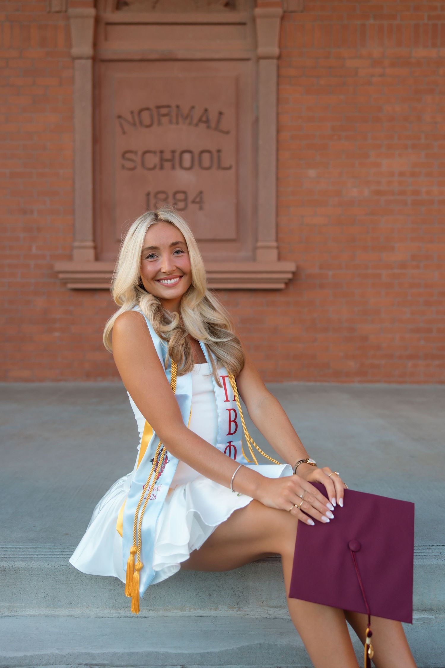 Smiling blonde graduate poses in white dress with Pi Beta Phi stole and maroon cap at Normal School 1884 building.
