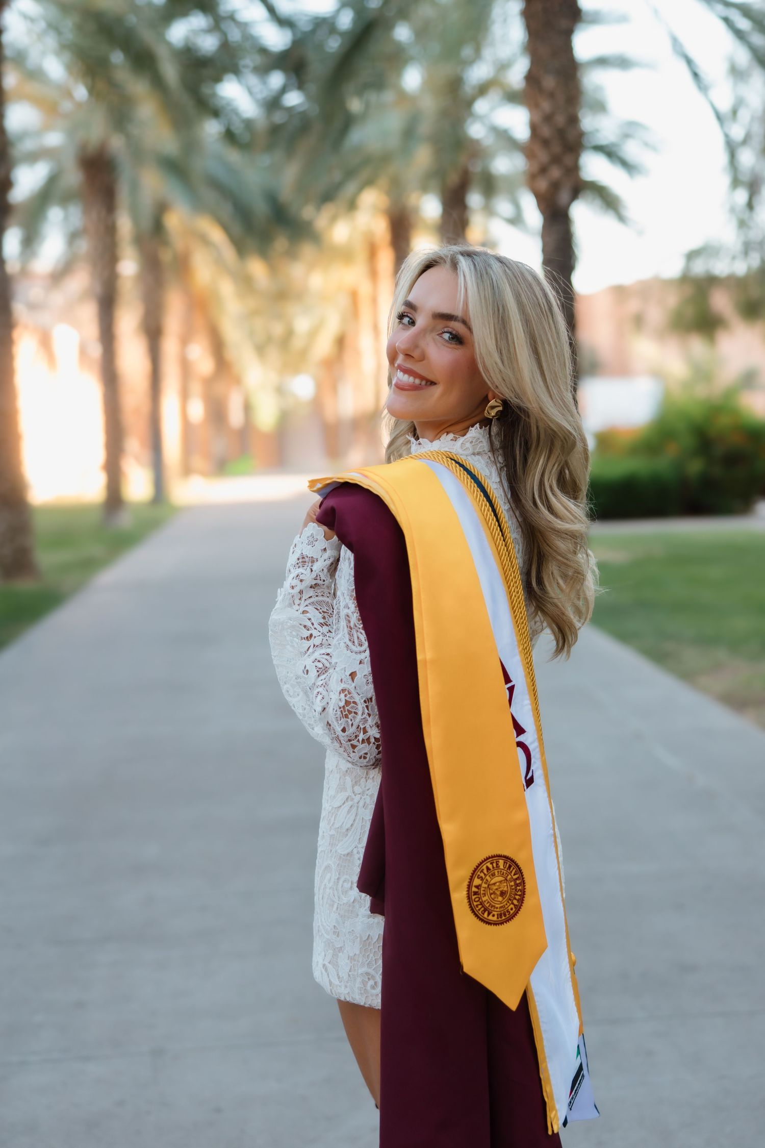 Smiling blonde graduate in white lace dress with maroon gown and gold stole on palm tree lined path.