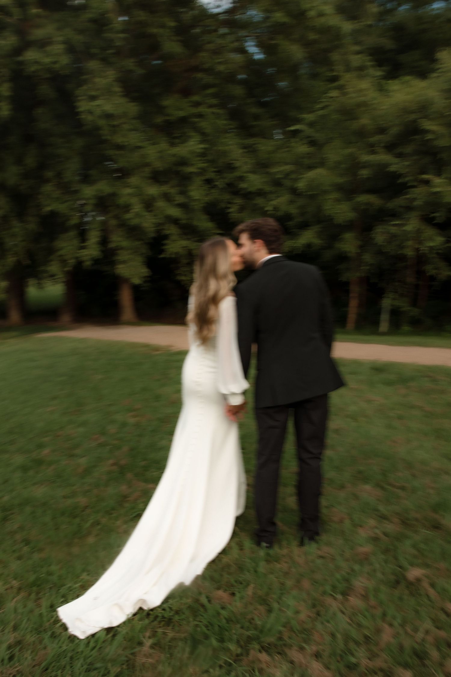 Bride and groom sharing a romantic kiss on a lush green lawn surrounded by trees.