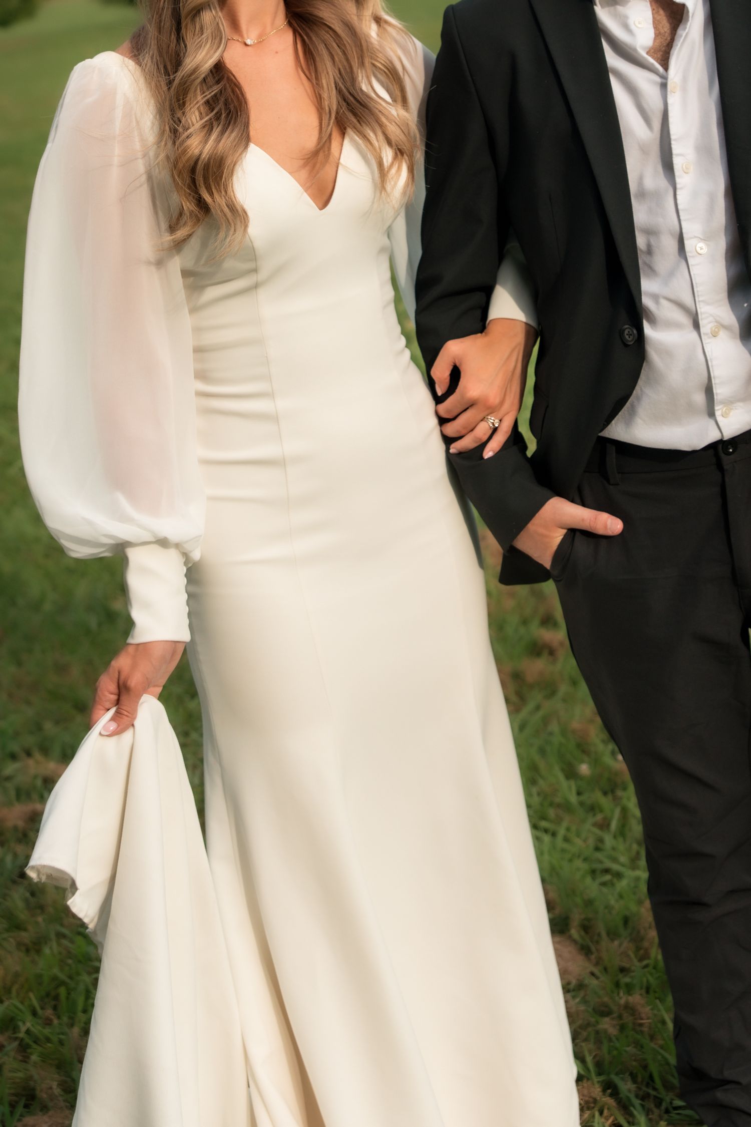 Bride in fitted white gown with sheer balloon sleeves holding hands with groom in black suit.