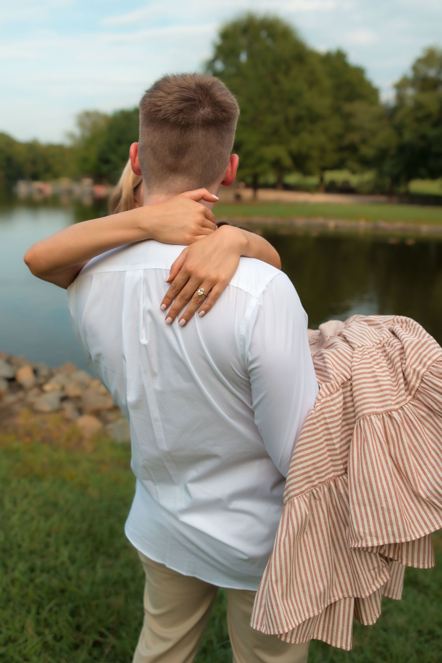 Couple embracing near a lake in a park, woman with ring wrapping arms around man in white shirt.