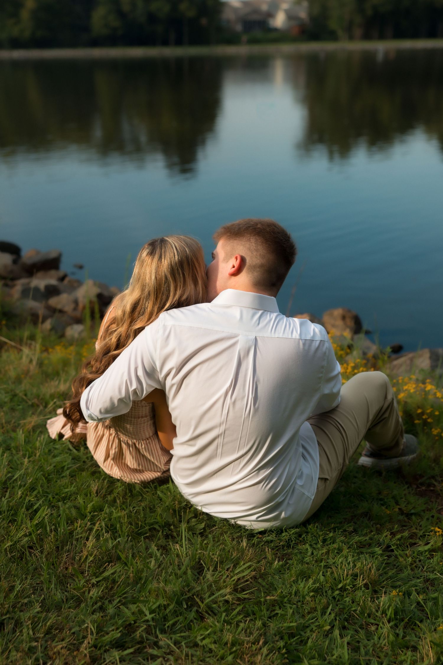 Couple sitting together on grass by a calm lake, embracing romantically at sunset in a serene outdoor setting.