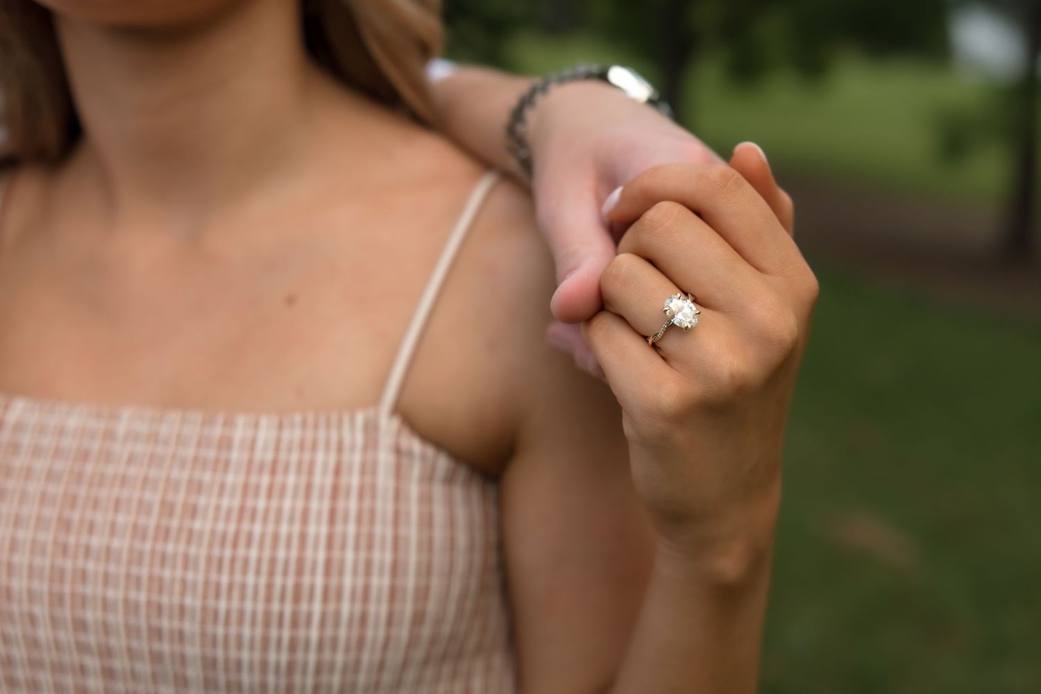 Close-up of a woman's hand displaying a diamond solitaire engagement ring while touching her shoulder outdoors.