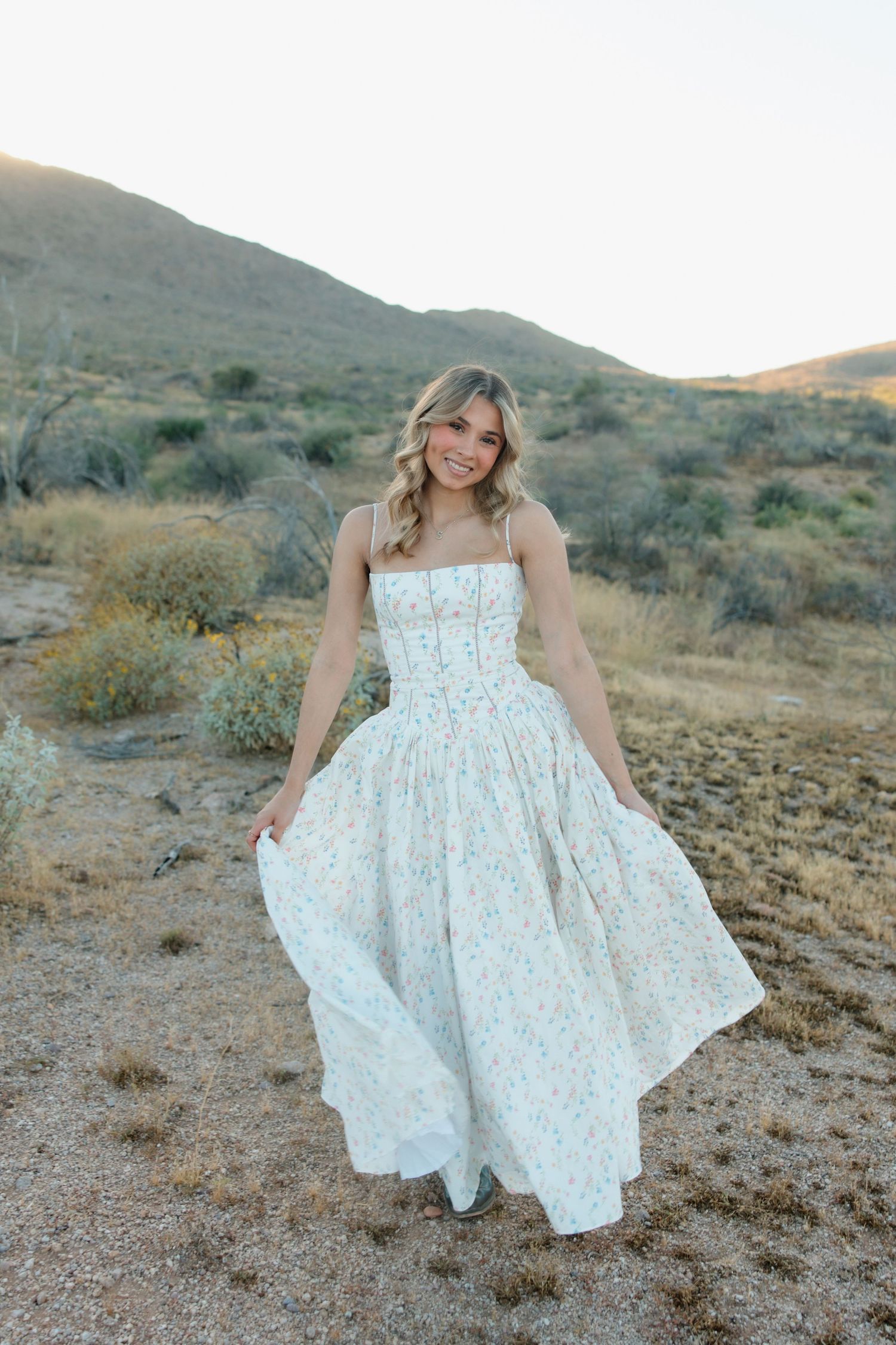 Young woman in floral maxi dress twirling in desert landscape with hills and shrubs at golden hour.