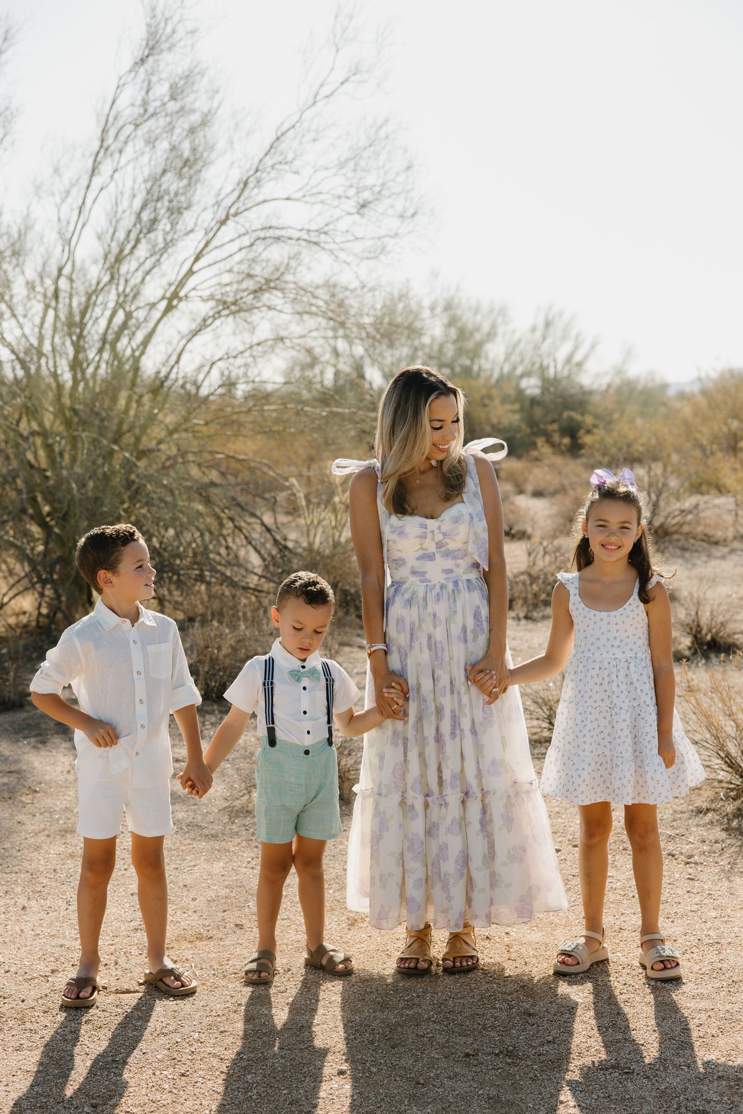 A woman in a floral maxi dress holds hands with three children in a desert landscape.
