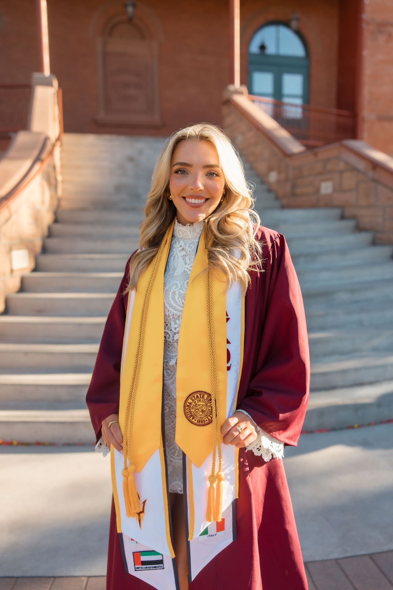 Smiling female graduate in maroon cap and gown with gold stole poses on university steps in sunny outdoor setting.
