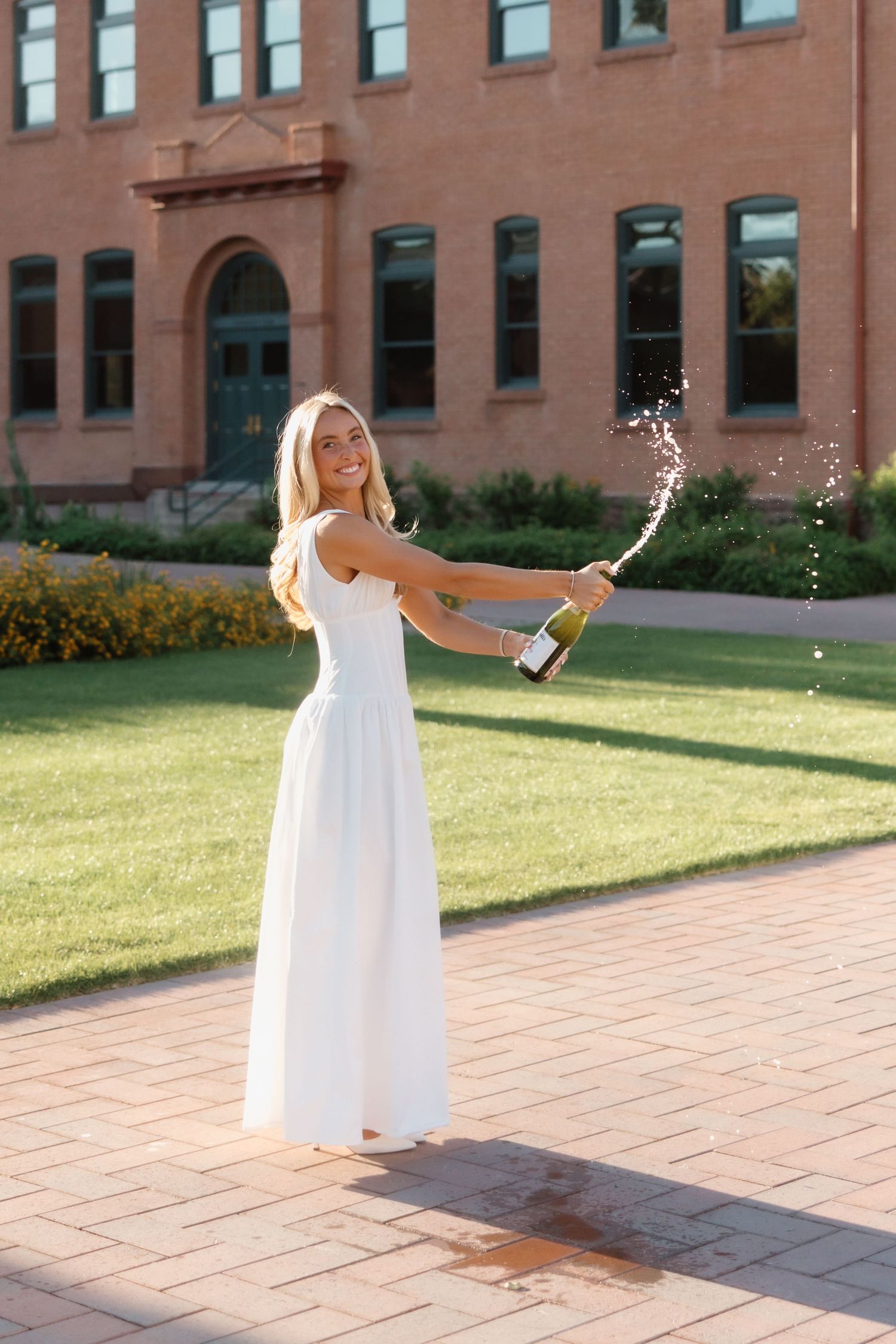 Smiling blonde woman in white dress popping champagne bottle outdoors near brick building on sunny day.