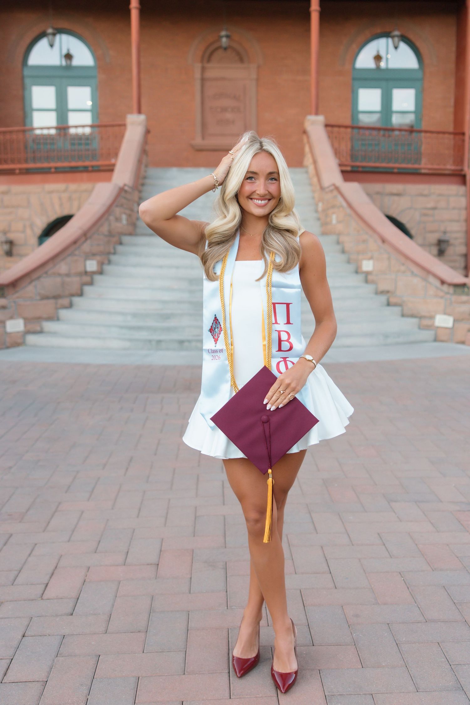 Smiling blonde woman in white dress with Pi Beta Phi graduation stole holding maroon cap on brick plaza.