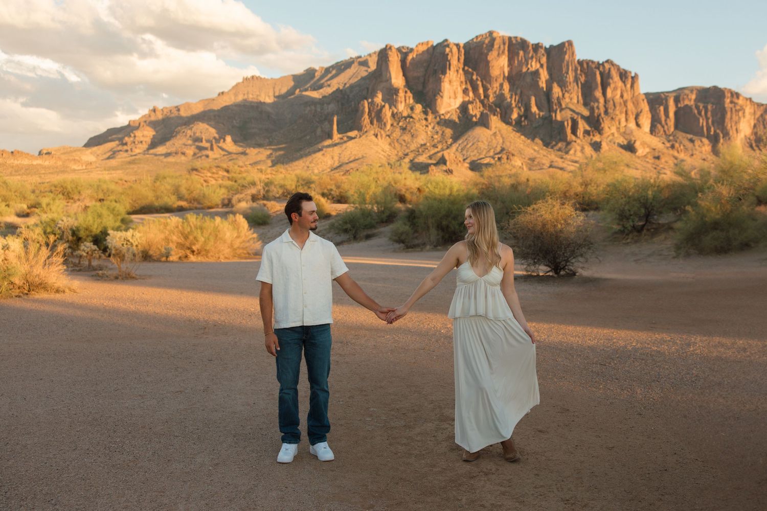 Couple holding hands in Arizona desert with Superstition Mountains at golden hour, wearing white outfits.