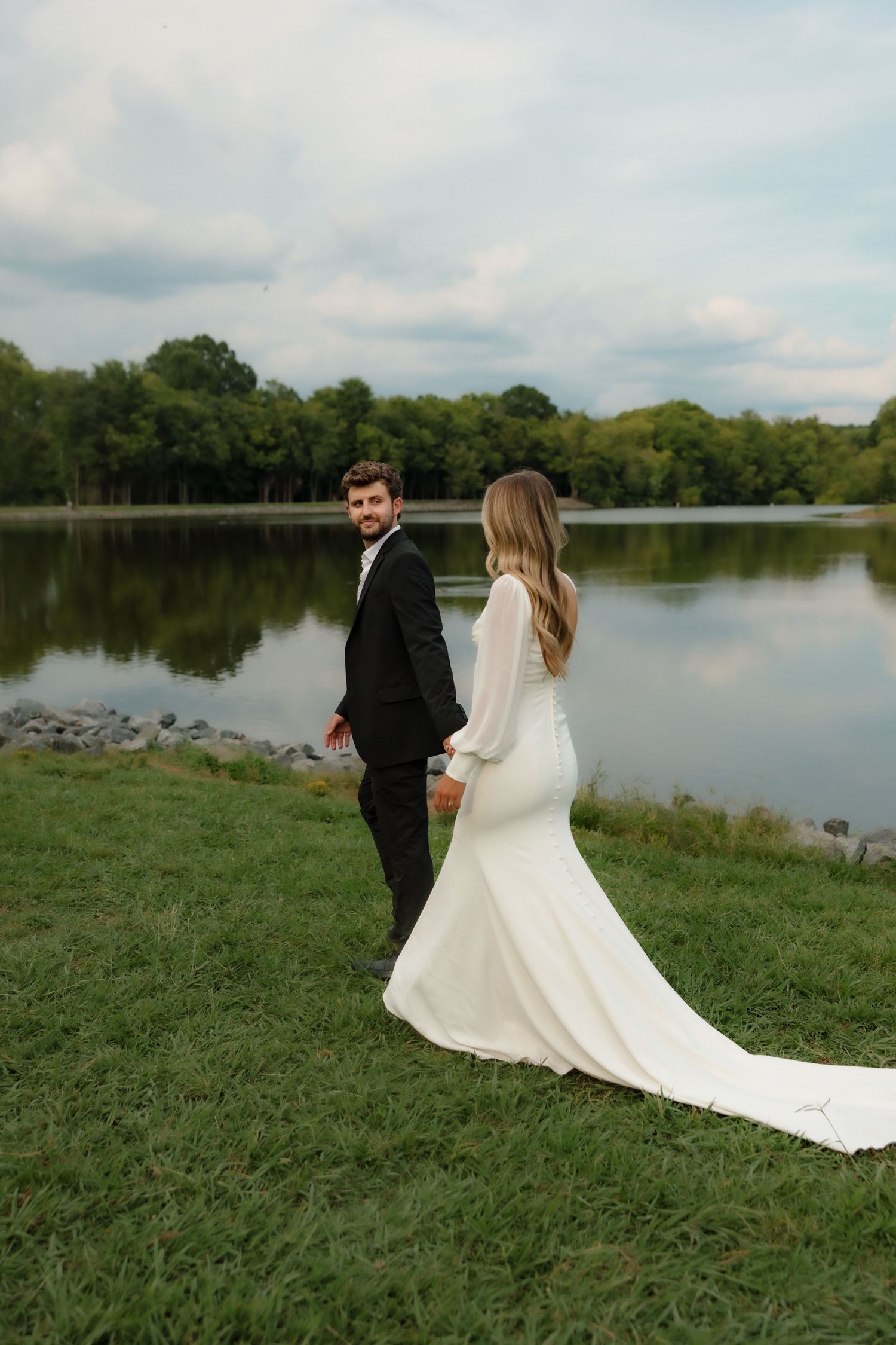 Wedding couple holding hands by a reflective lake, groom glancing back in black suit and bride in white gown.