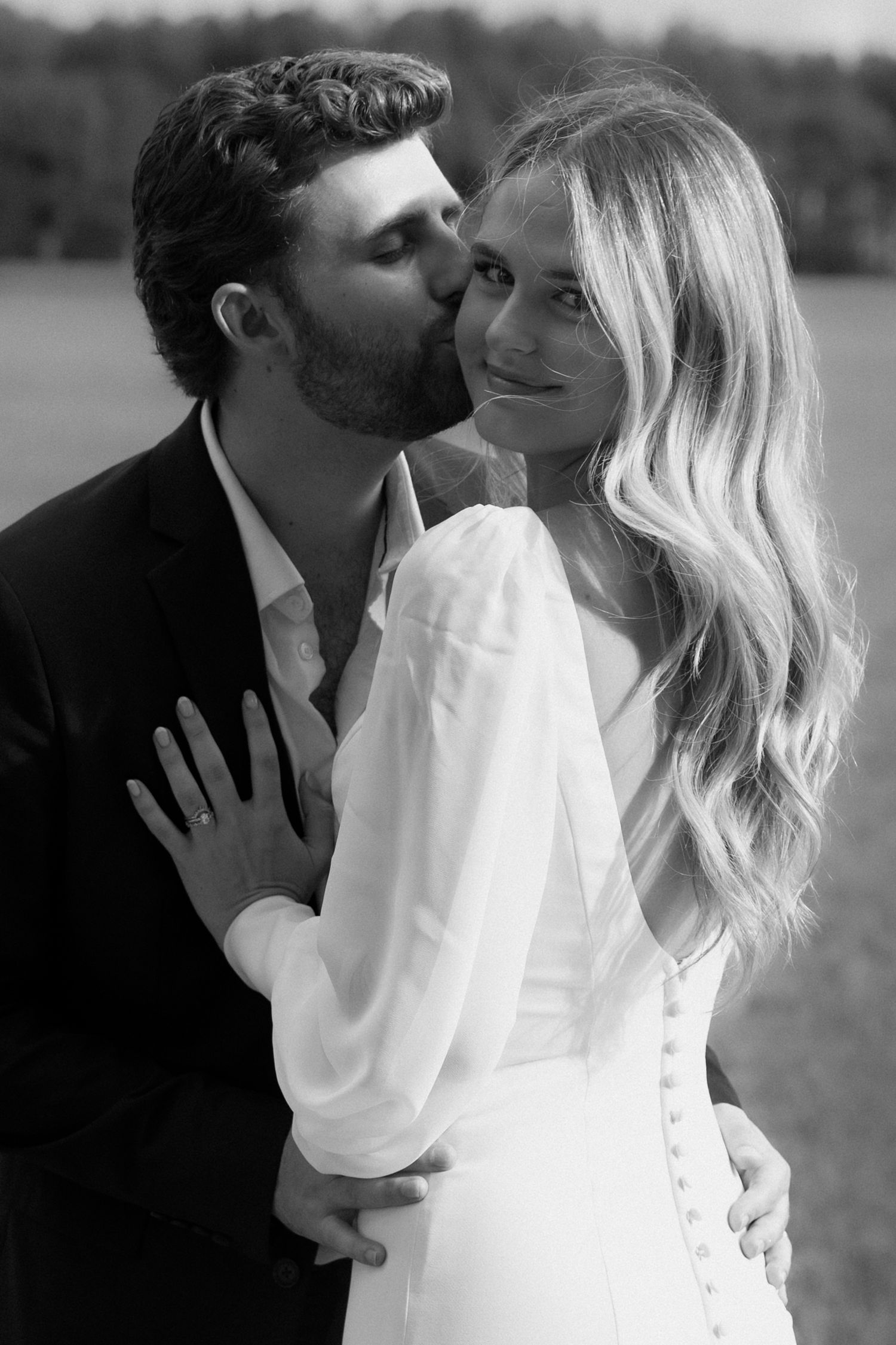 Black and white photo of a groom kissing bride on cheek, she smiles in white dress outdoors.